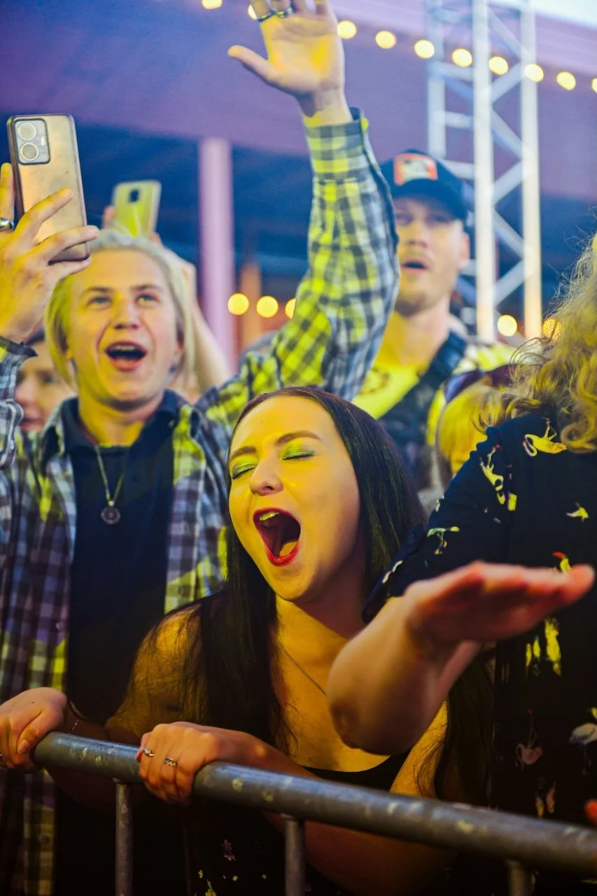 Group of young people at a live music event, displaying excitement and enjoying music, with some taking photos or videos and others singing or dancing.