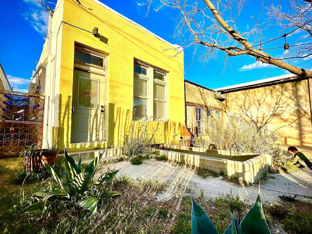 Front yard garden of a yellow house with a door and windows, some plants, a tree with no leaves, and shadows cast by the tree and house against a blue sky.