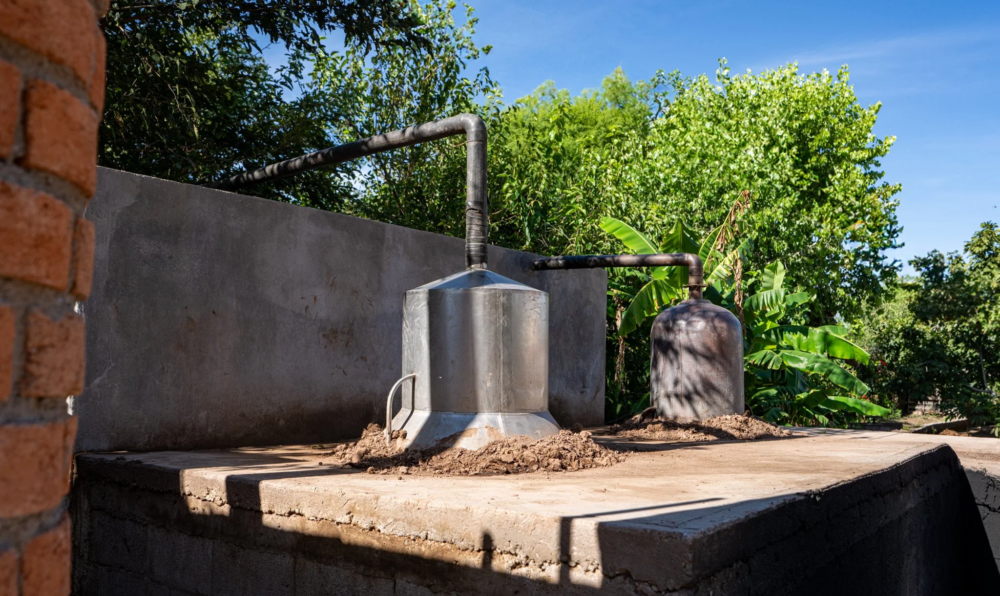 Metal rainwater tanks with pipes on a concrete platform in an outdoor garden area with trees and blue sky