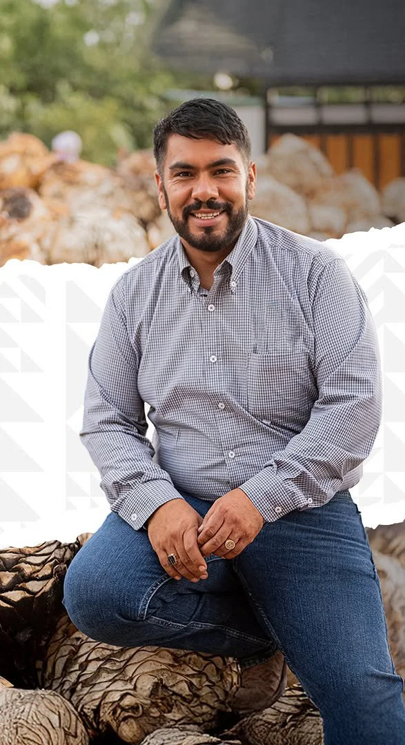 A man with dark hair and a beard, smiling, wearing a light checkered shirt and jeans, sitting outdoors on a large piece of wood with rocks and trees in the background.