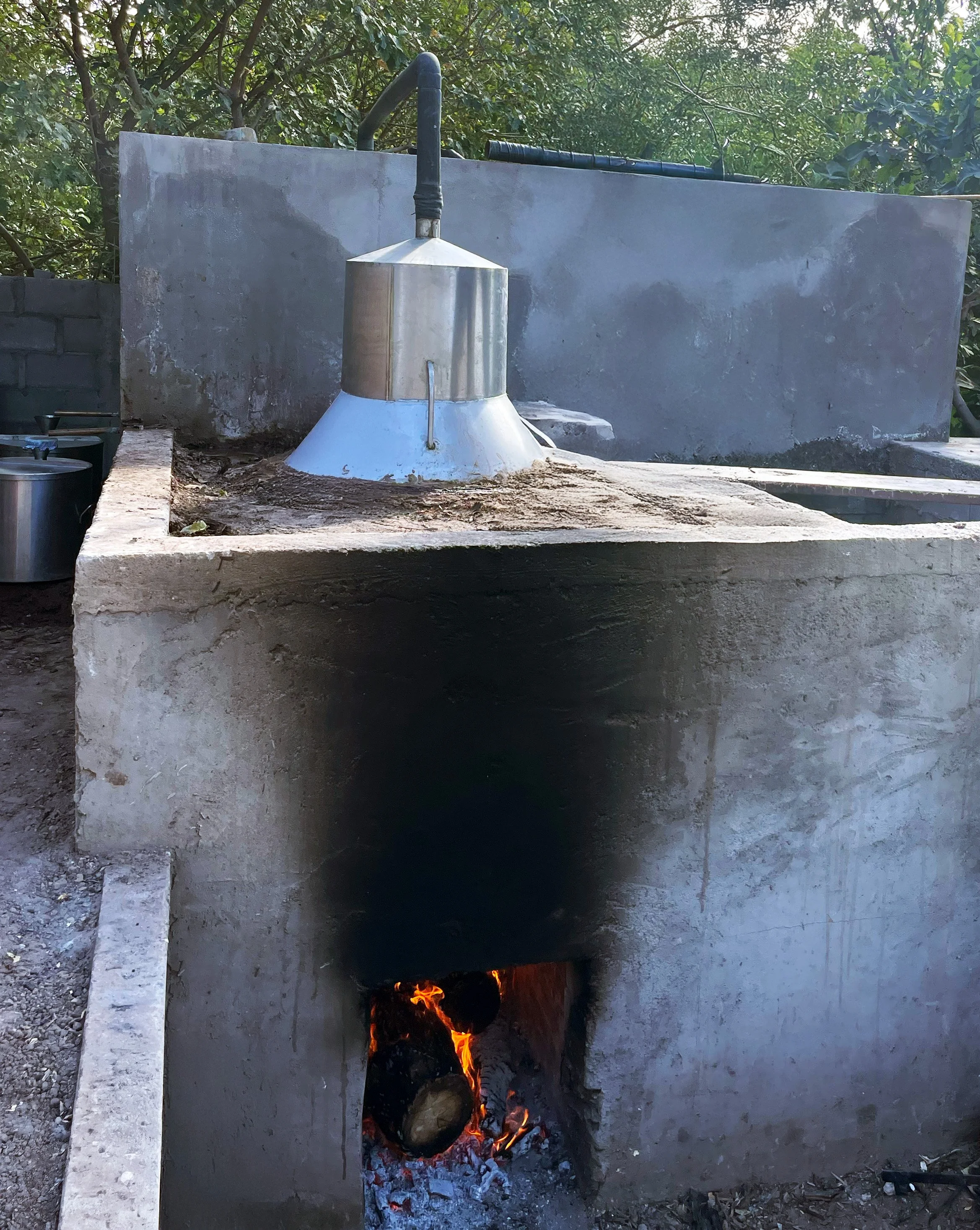 A traditional outdoor wood-fired brick oven with a metal exhaust pipe, burning logs inside, and a concrete chimney or divider behind it.