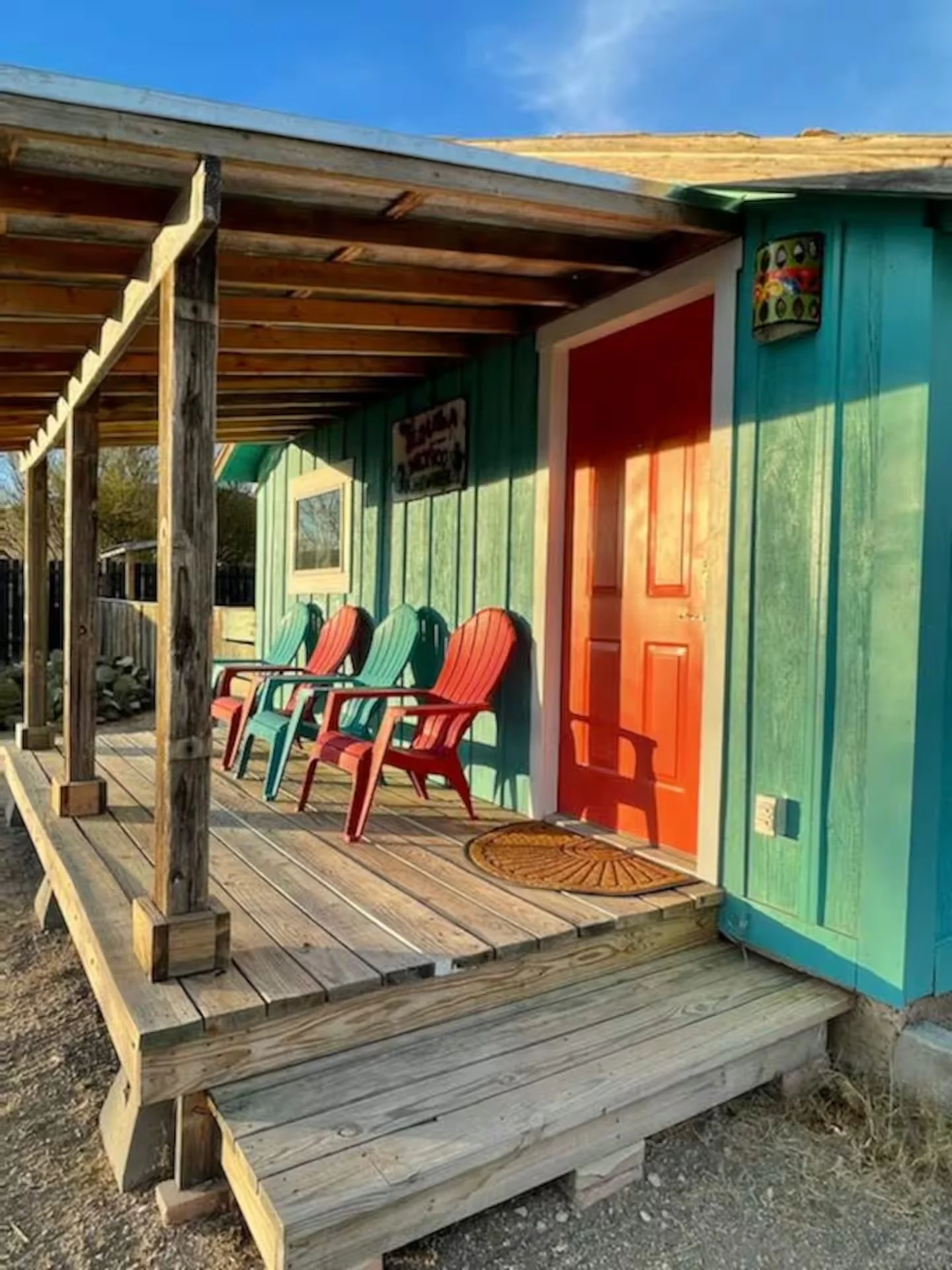 Colorful small house with a wooden porch, two Adirondack chairs (one red, one green), a red door, and a decorative wall piece, under a partially covered roof, with a clear blue sky in the background.