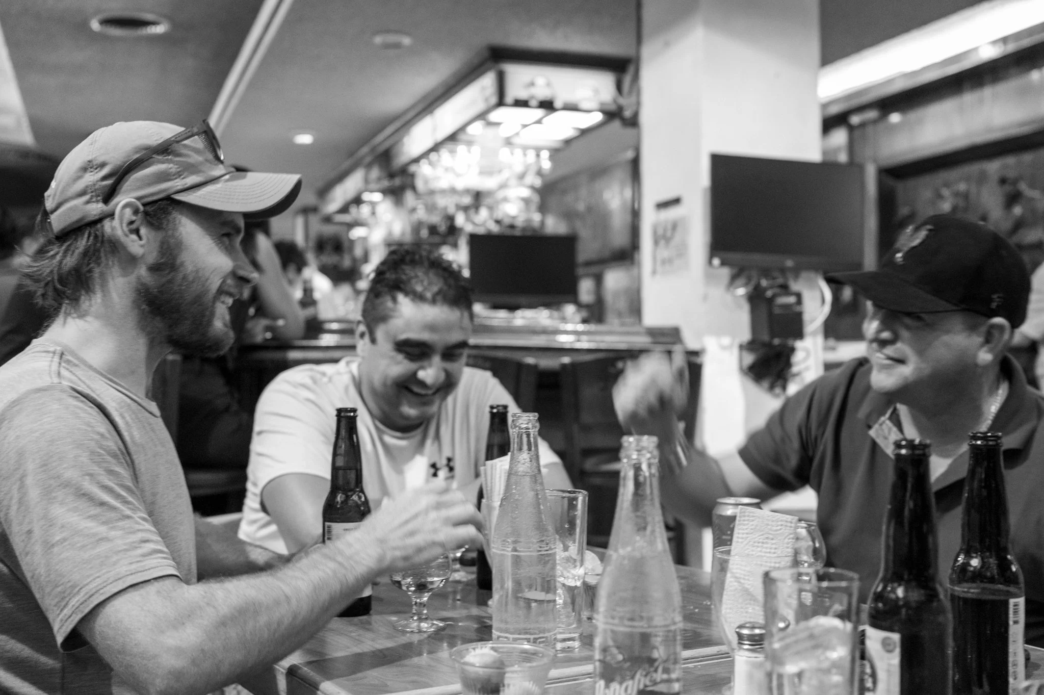 Three men sitting at a bar counter, smiling and talking, with beer bottles and glasses in front of them, in a lively, casual environment.