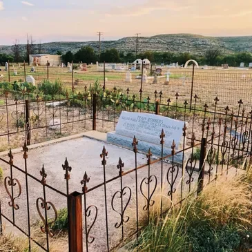 A fenced grave site with a blue headstone in a rural cemetery, with open land and hills in the background.