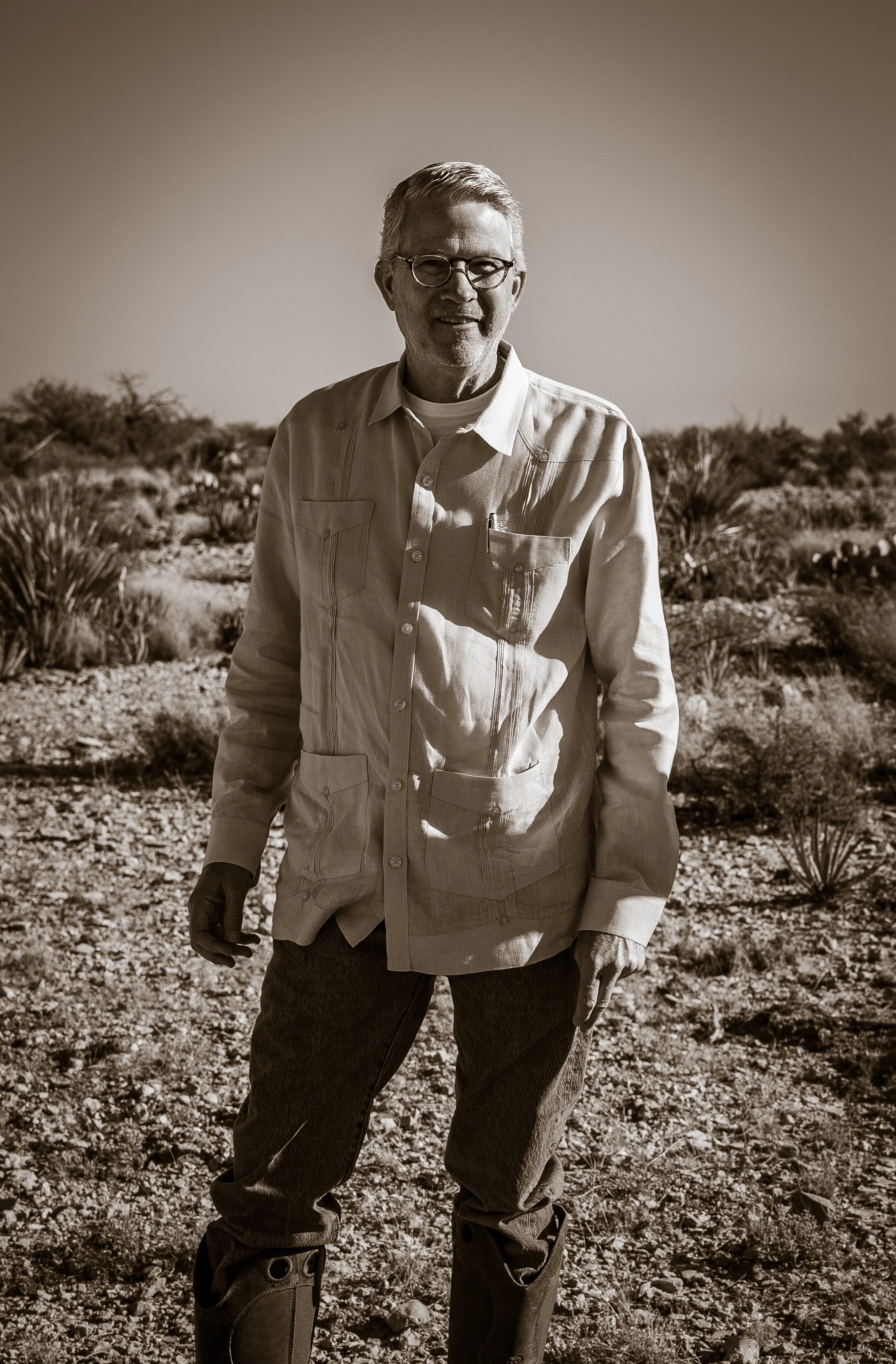 Tall Texan Distilling founder, Richard Lonquist, on his ranch in Sanderson, Texas in the Chihuahuan Desert is wearing glasses, a long-sleeved button-up shirt, and rubber boots, with sparse vegetation around him.