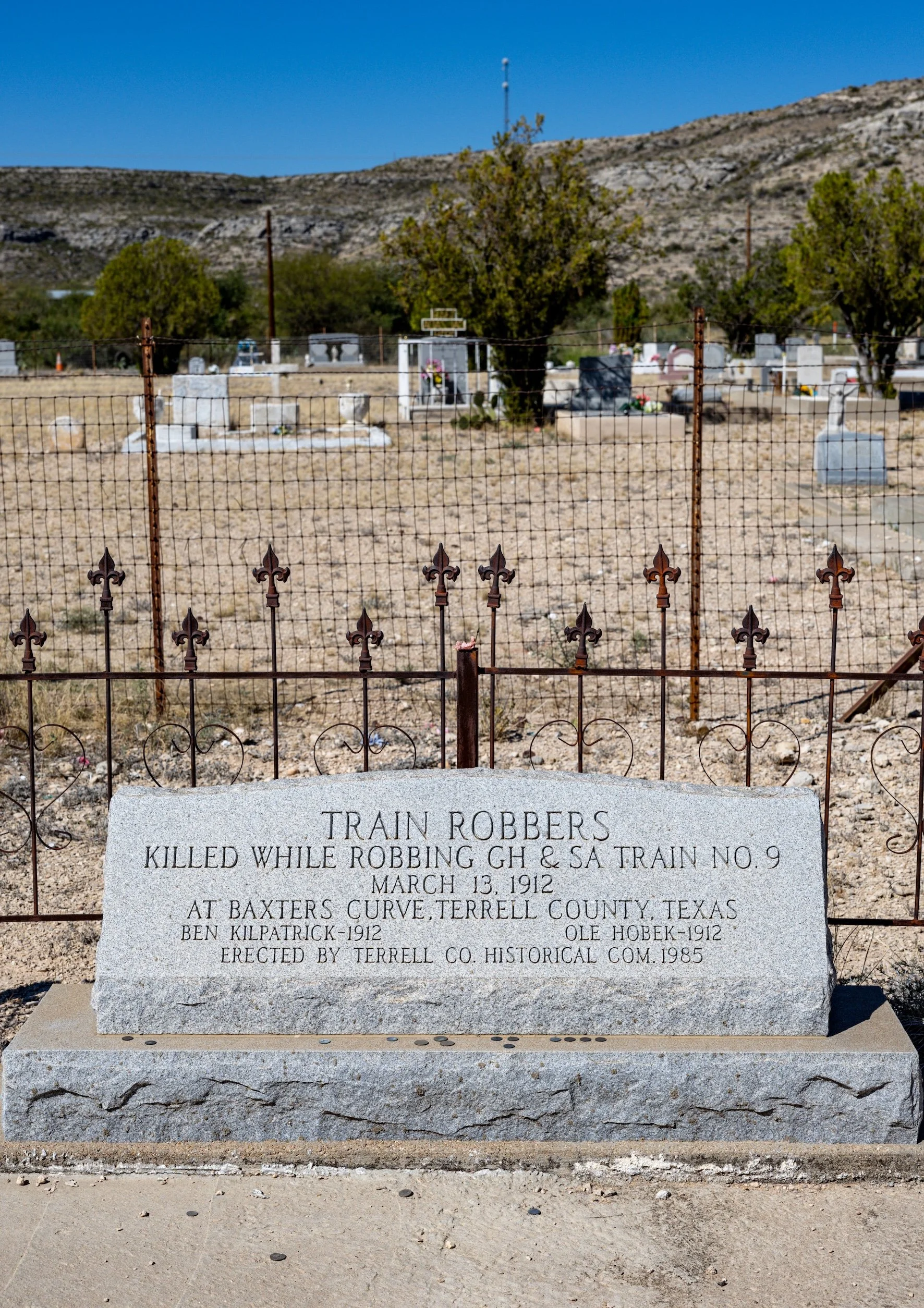 A grave site with a stone marker for train robbers, located at Baxters Curve, Terrell County, Texas. The marker commemorates train robbers killed during a robbery on March 13, 1912, with names and details inscribed. The area is fenced, with a dry landscape, a few trees, and hills in the background under a bright blue sky.