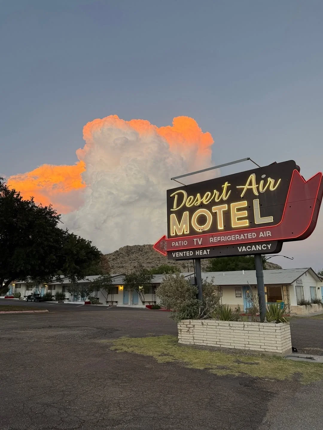 Neon sign for Desert Air Motel with an arrow pointing left, showing amenities such as patio, TV, refrigerated air, vented heat, and vacancy, with a cloudy sky and mountains in the background.