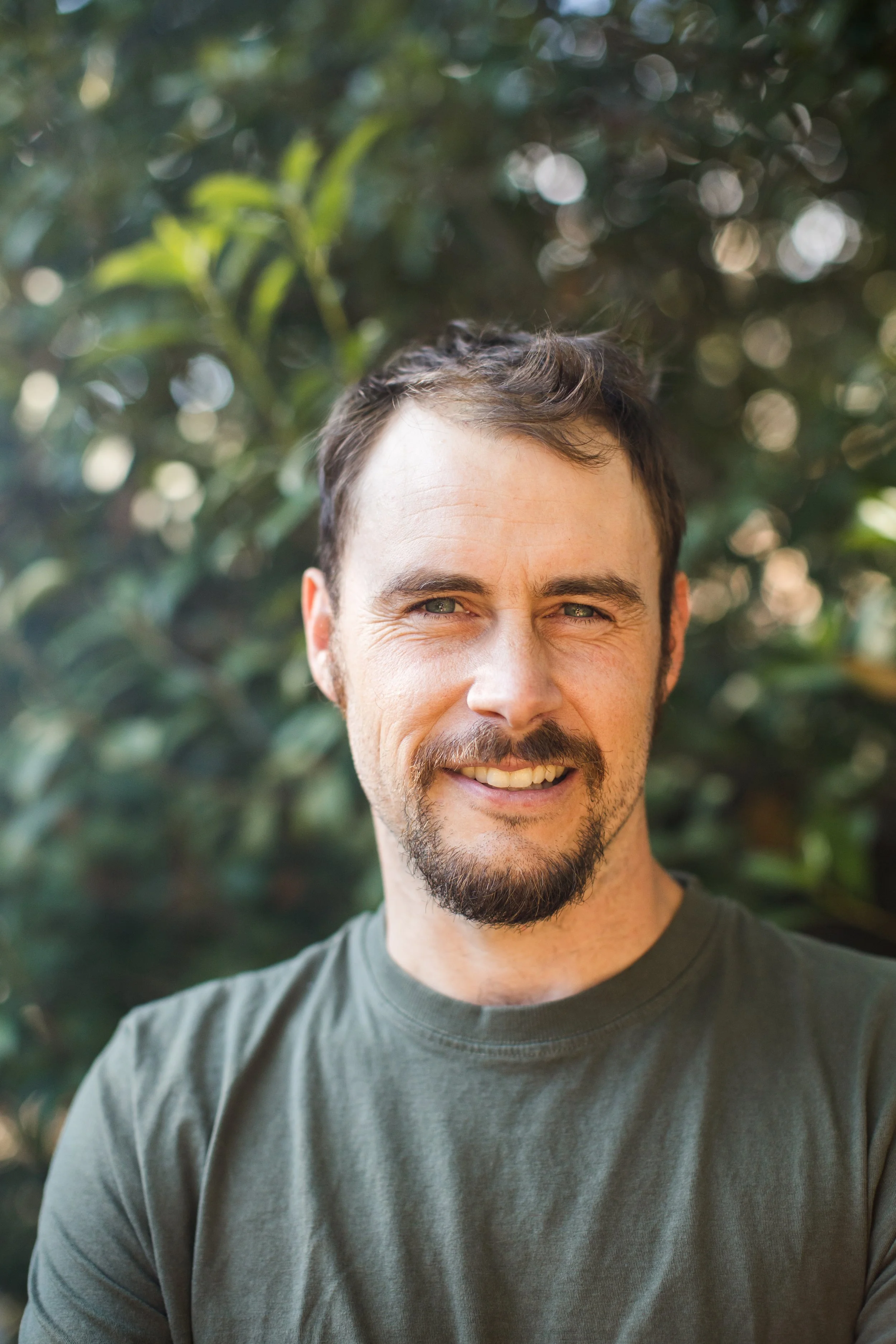 A smiling man with dark hair, a beard, and a mustache, wearing a green T-shirt, outdoors with blurred greenery in the background.