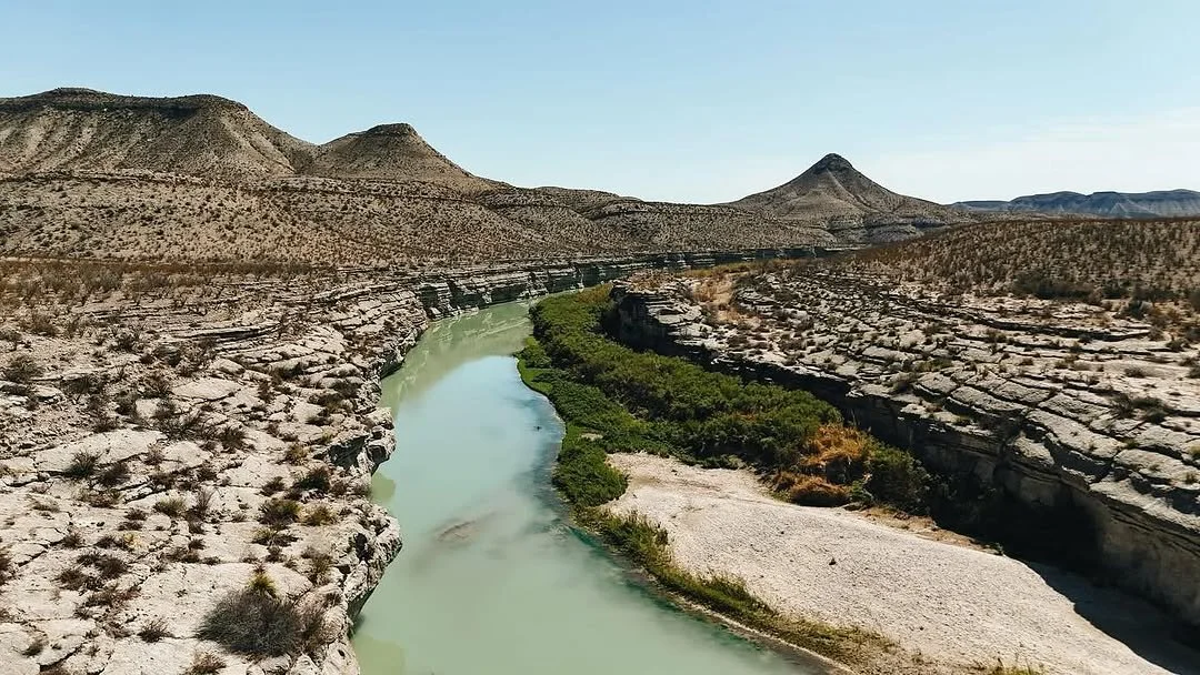 A winding river running through a rocky desert landscape, with sparse vegetation and distant hills under a clear sky.