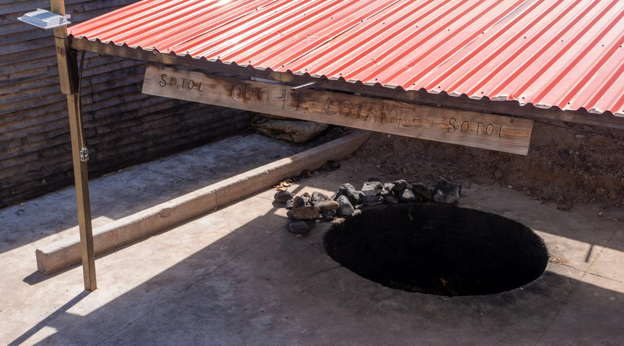 An earthen pit oven with rocks piled beside it, underneath a slanted metal roof with a wooden sign that says "SOTOL ORO DE COYAME".