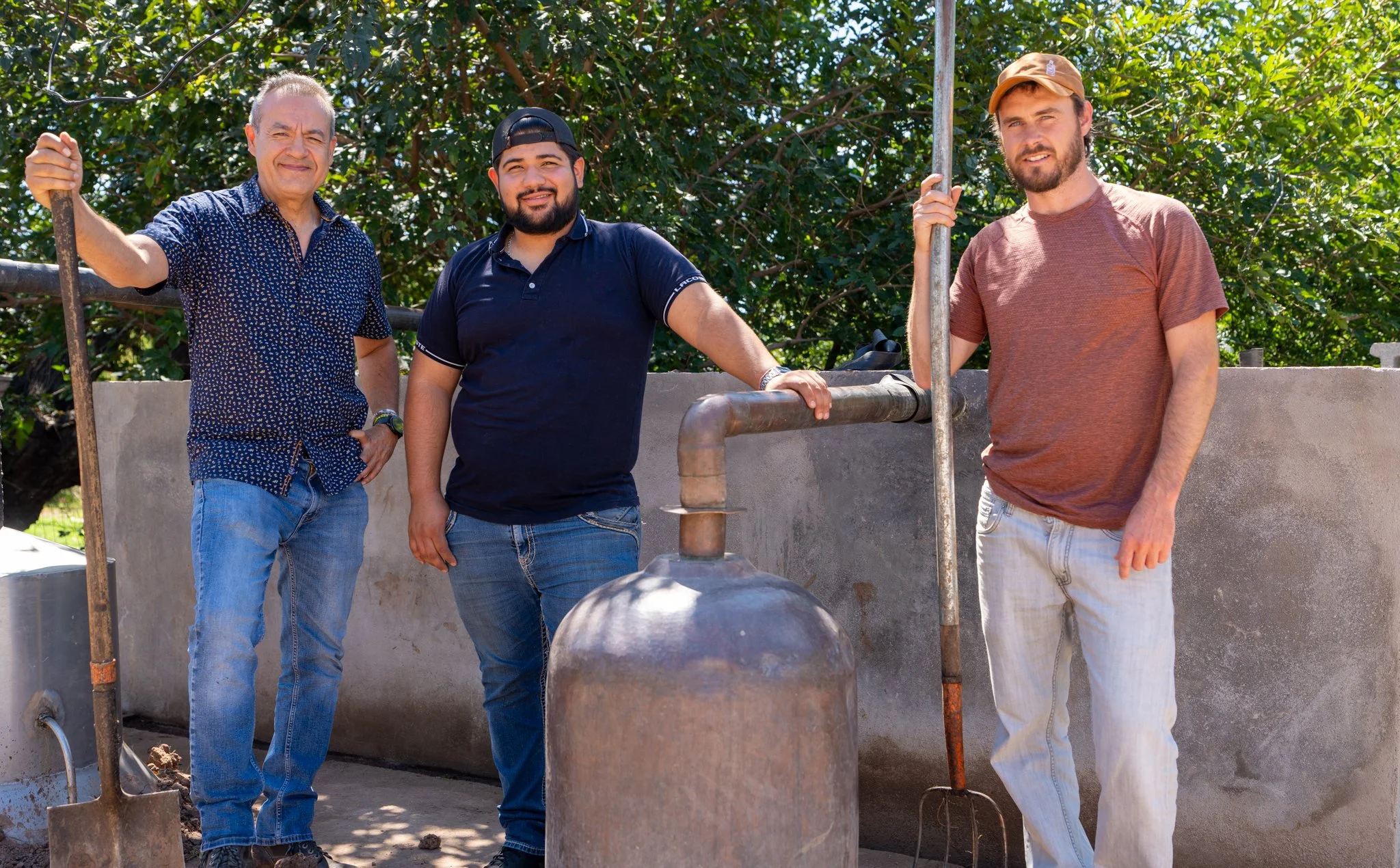 Three men standing outside near pipes and construction tools, smiling at the camera, with trees and a concrete wall in the background.