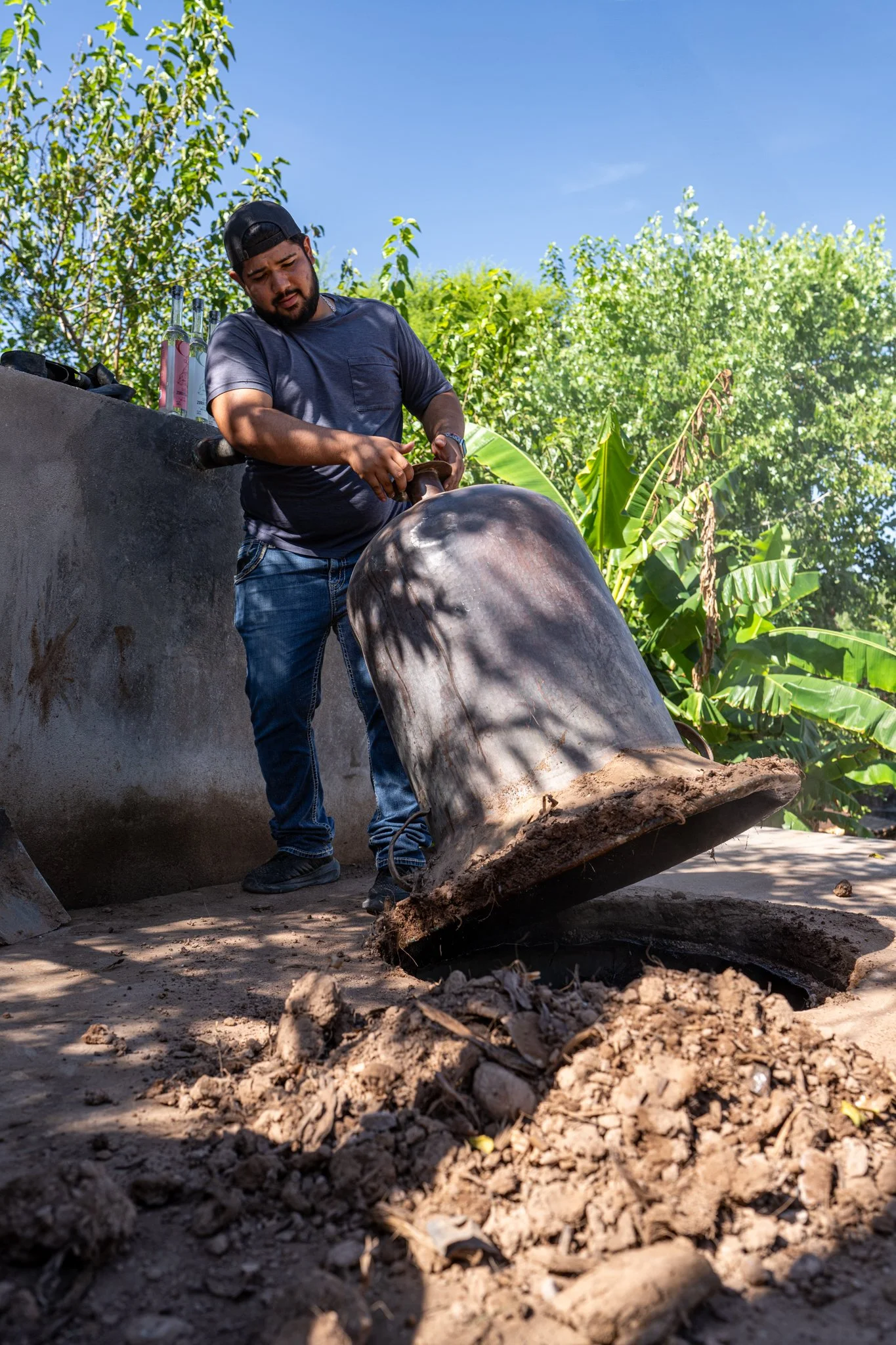 A man in casual clothes and a black baseball cap is using a tool to extract something from an underground oven.