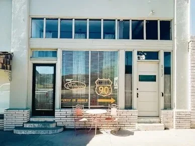 Shopfront with large windows and a door, with a display inside showing a '90' sign, and two small tables with chairs outside.