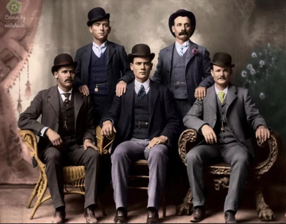 Vintage photograph of six men dressed in early 20th century suits and hats, posing for a formal portrait with two seated on ornate chairs and four standing behind them.