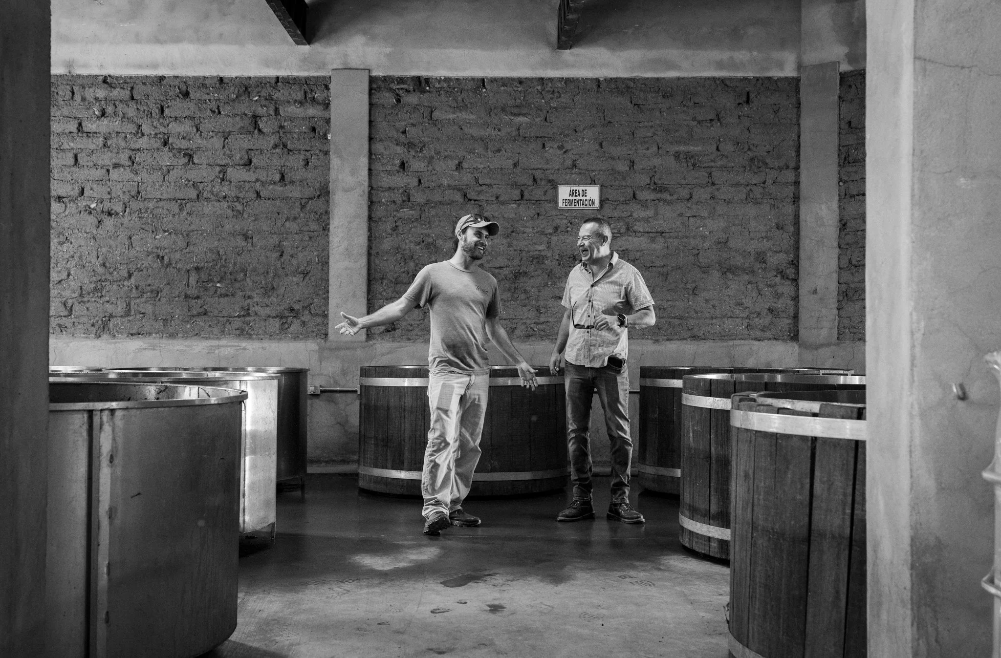 Two men are smiling and talking inside a brewery or winery, surrounded by large wooden fermentation tanks. The background includes a brick wall and a sign that reads 'Area de Fermentacion'.