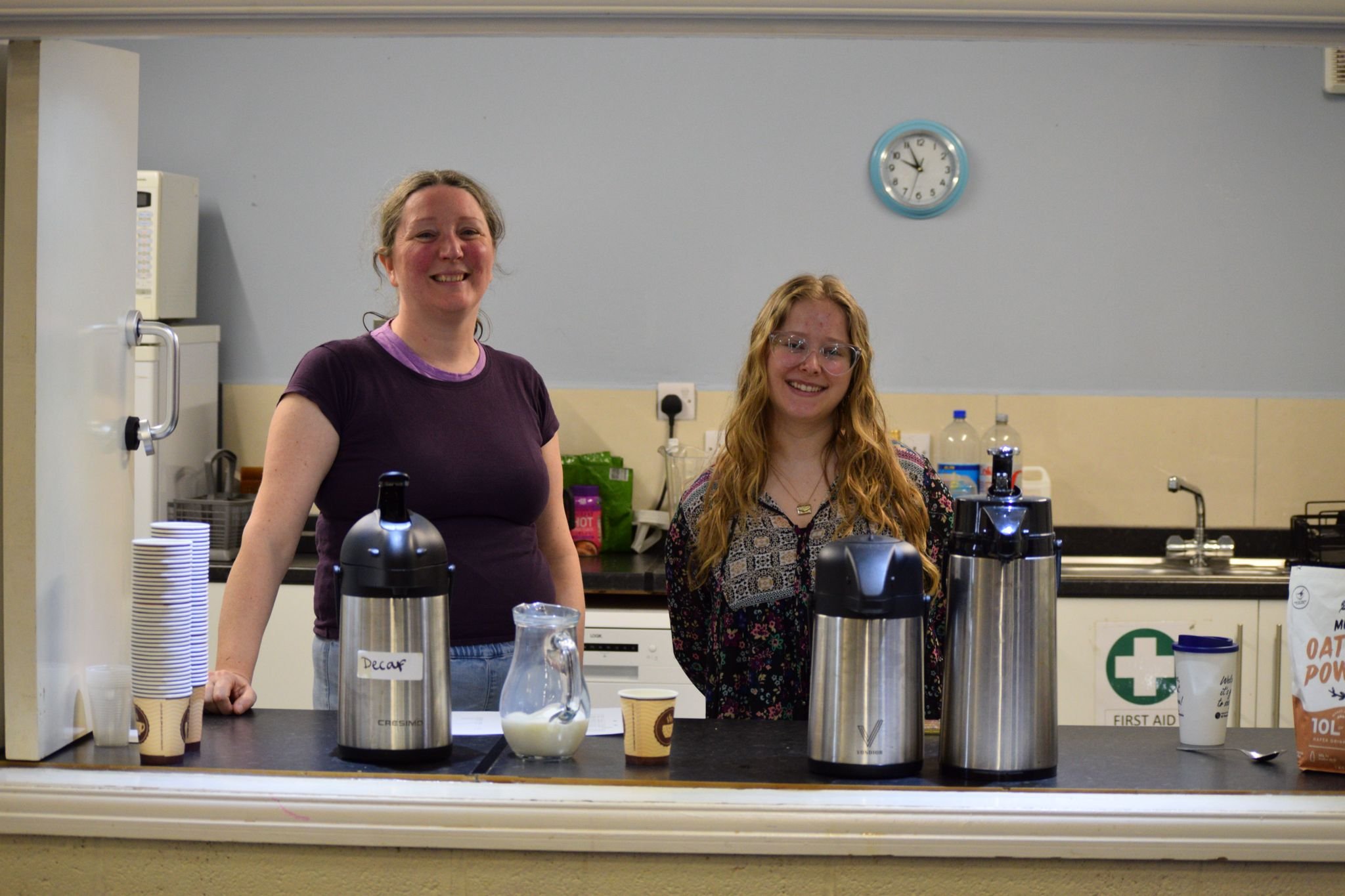 Two smiling women standing behind a counter in a kitchen or break room. There are three large thermal coffee dispensers, a jug of milk, paper cups, and a bag of oats on the counter. A clock on the wall shows 10:09.