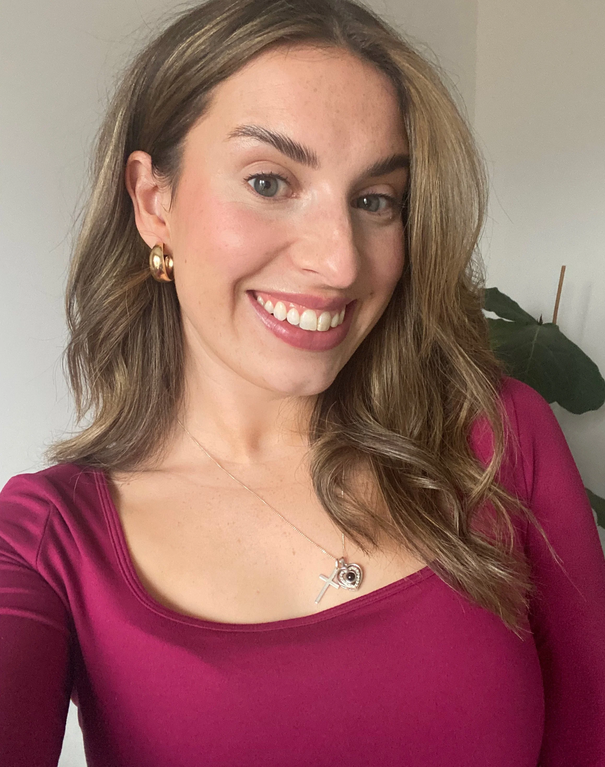 A woman with long dark hair, blue eyes, and fair skin, smiling and wearing a black top, earrings, and a cross necklace, standing indoors near a staircase.
