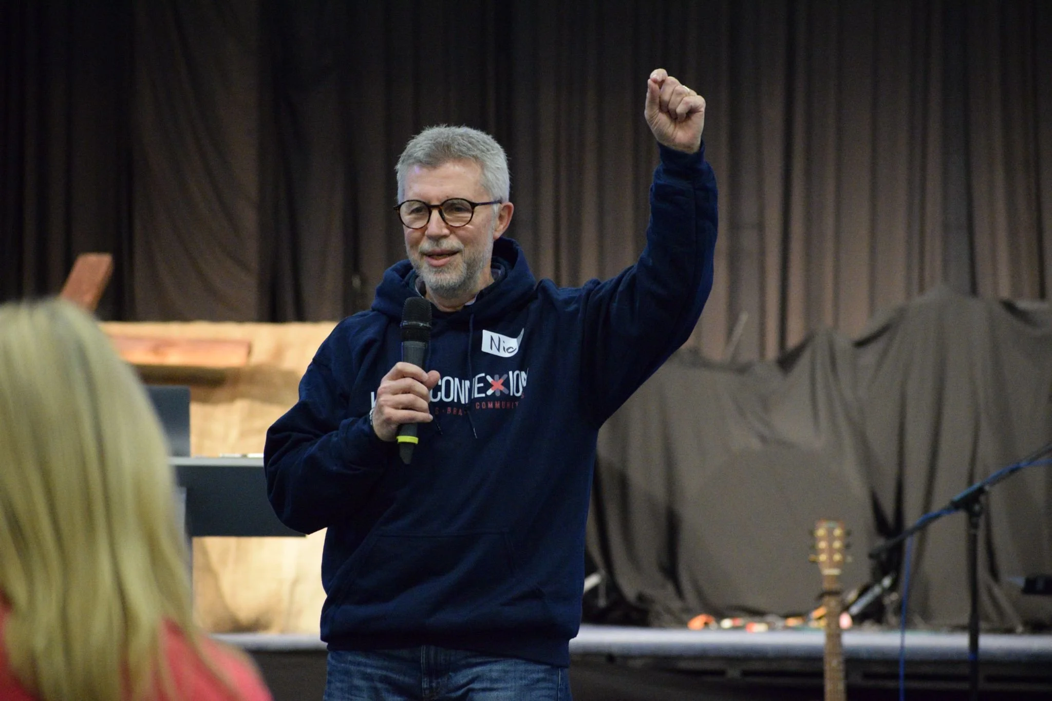 A man with gray hair, glasses, and a beard is speaking into a microphone and raising his right fist. He is wearing a navy hoodie with a logo on the front, standing in front of a stage with dark curtains.