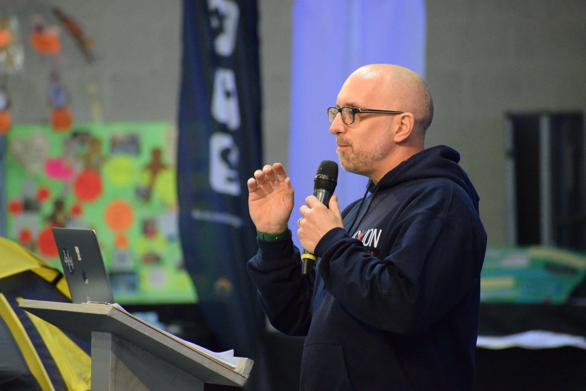 A man wearing glasses and a dark hoodie speaking into a microphone at a podium with a laptop, in front of a colorful background with a poster and a flag.