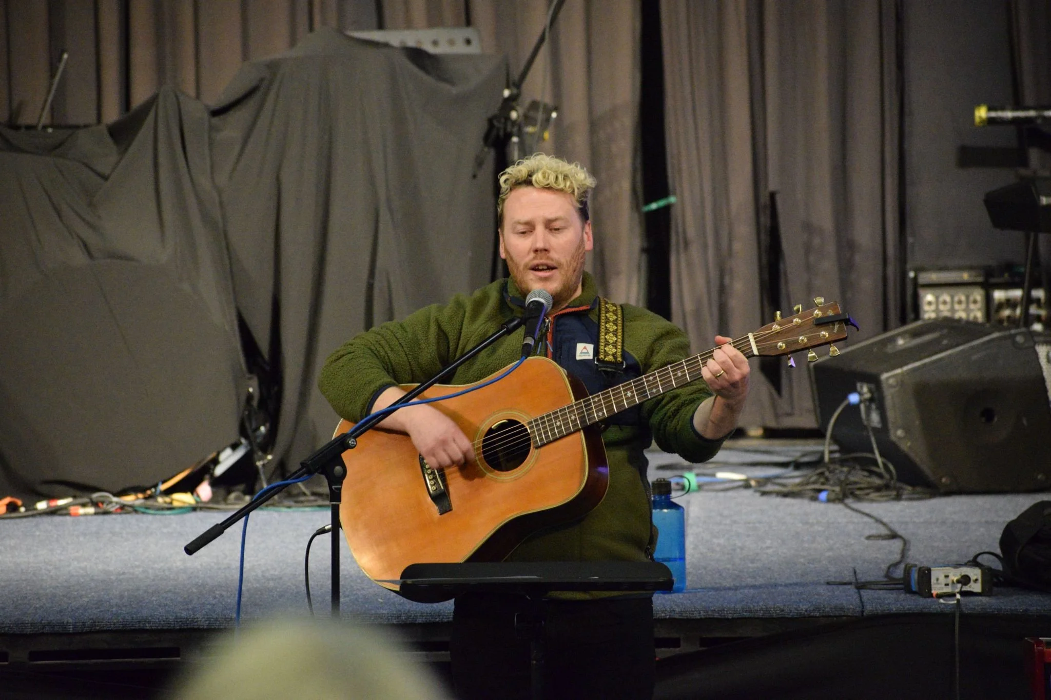 A man with blonde curly hair and a beard playing acoustic guitar and singing into a microphone on a stage with dark curtains and equipment in the background.