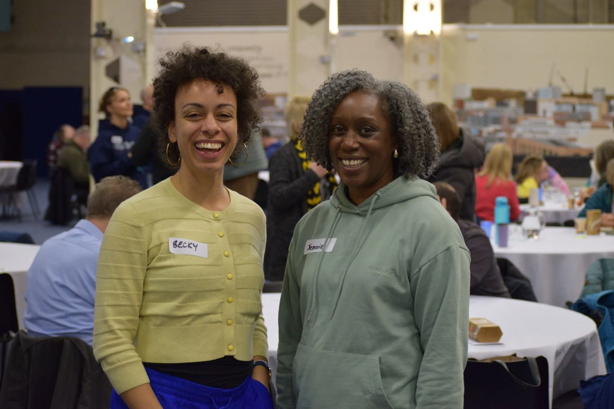 Two women standing together at a social event, smiling at the camera. They are wearing casual clothing, with name tags labeled 'Becky' and 'Jennie'. In the background, there are several people, tables, and a large model of buildings.
