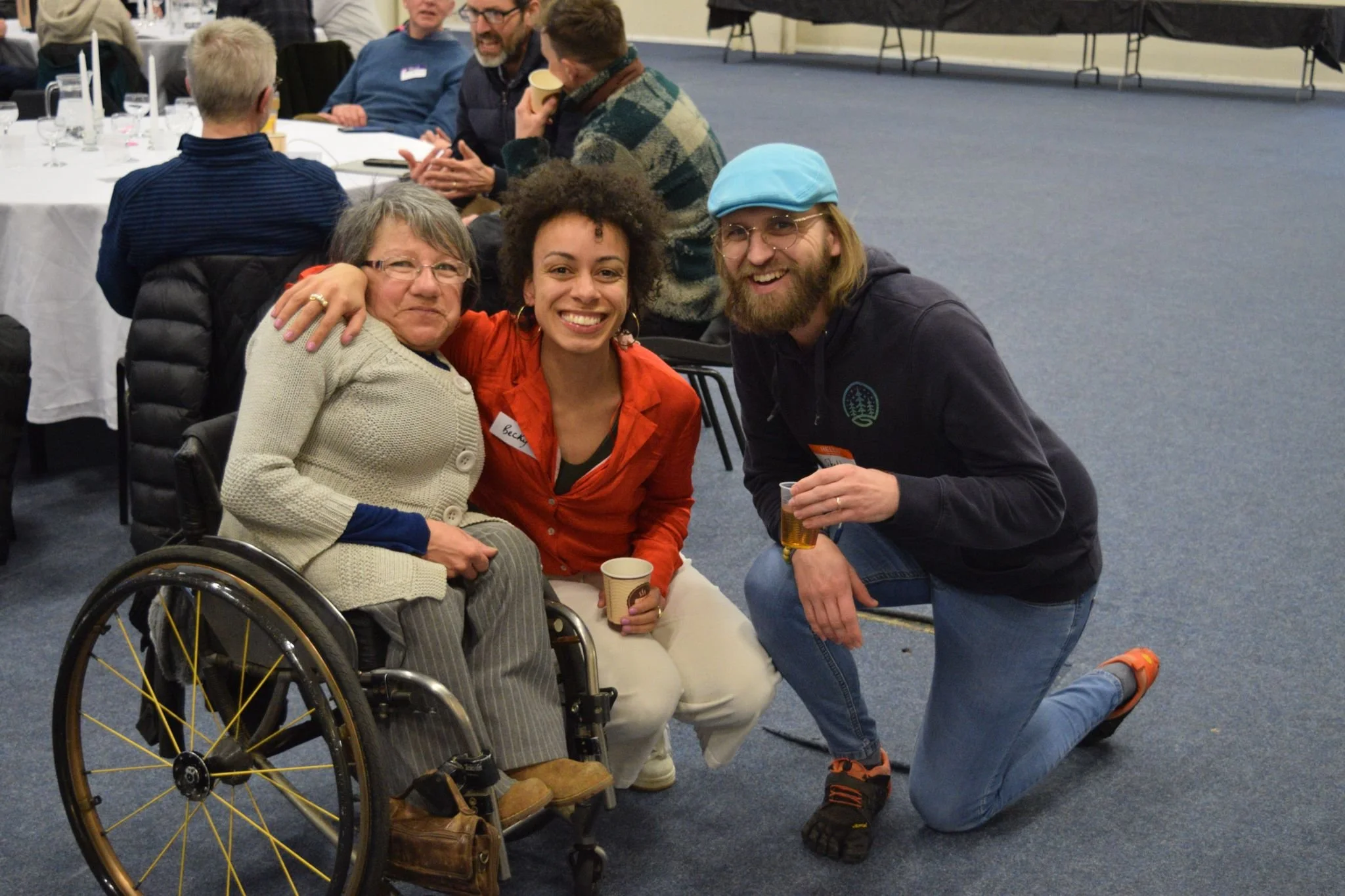 Three people smiling at a social gathering: an older woman in a wheelchair, a young woman with curly hair and a red jacket, and a young man with a beard, glasses, and a blue cap, holding drinks.