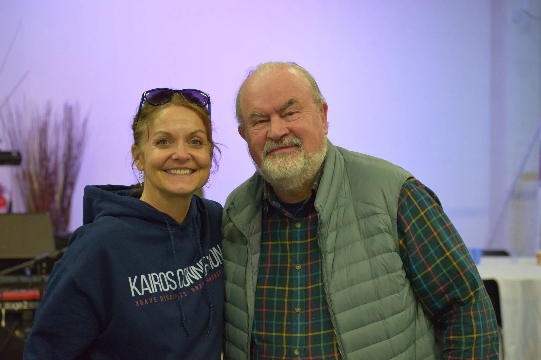 A woman with sunglasses on her head and a man with a beard smiling in an indoor setting.