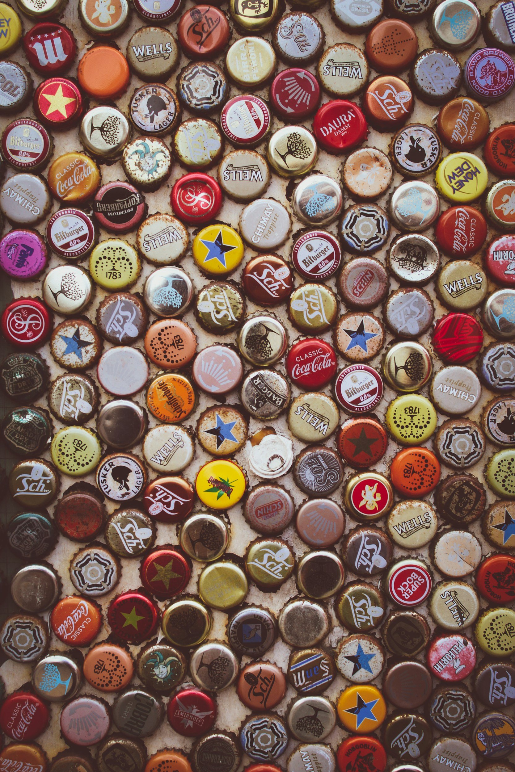 A collection of various soda bottle caps, including brands like Coca-Cola, Sprite, Dr. Pepper, and other craft and local brands, arranged closely on a surface.