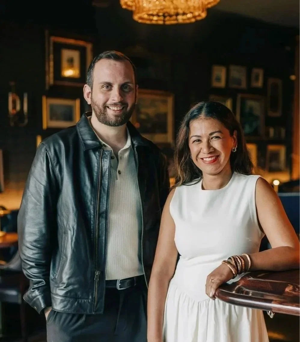 A man with a beard in a black leather jacket and a woman with dark hair in a white dress smiling in a dimly lit room with framed pictures on the wall.
