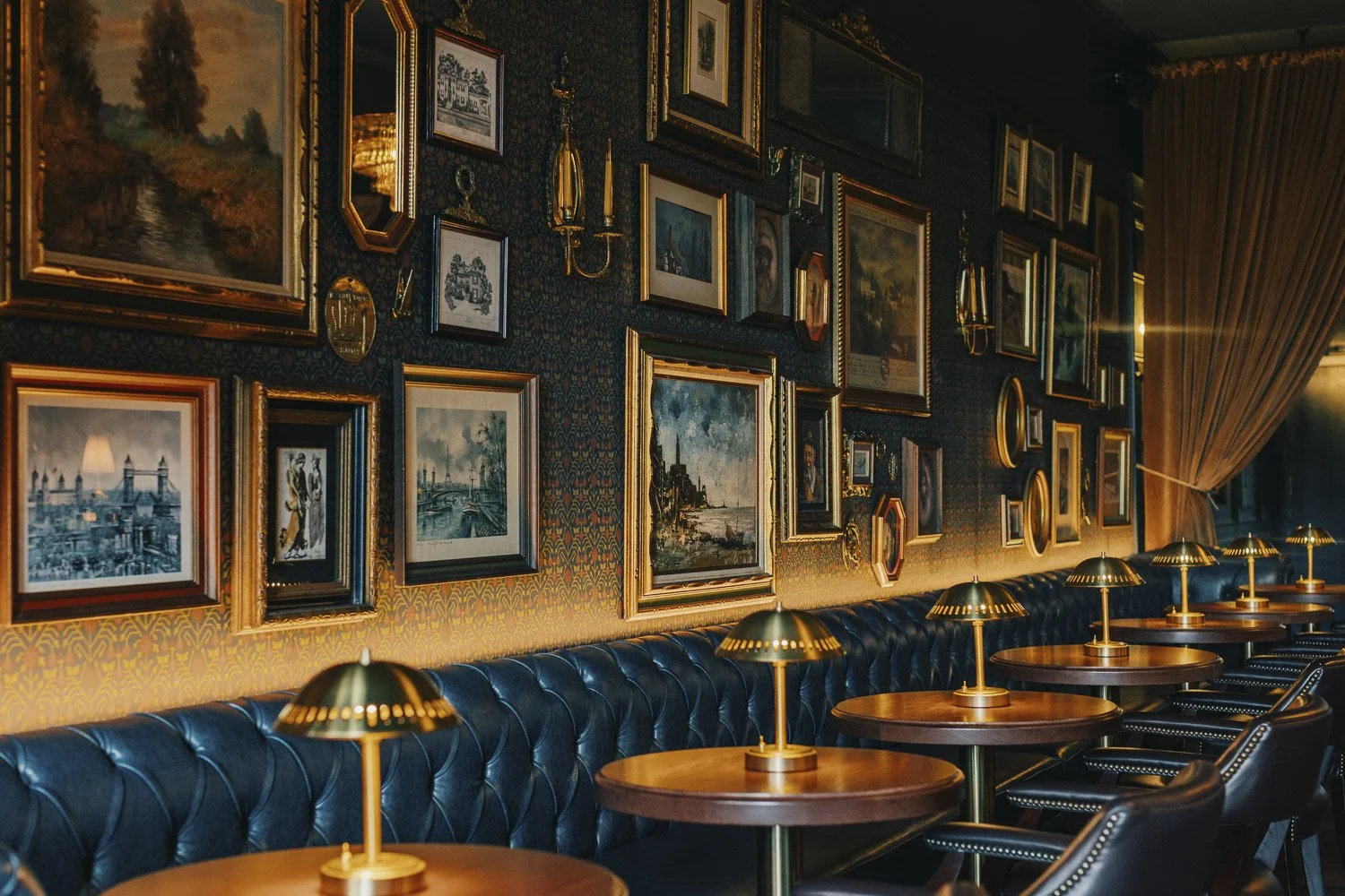 Gallery wall with vintage artwork on patterned wallpaper above chesterfield style banquette and dining tables with table lamps
