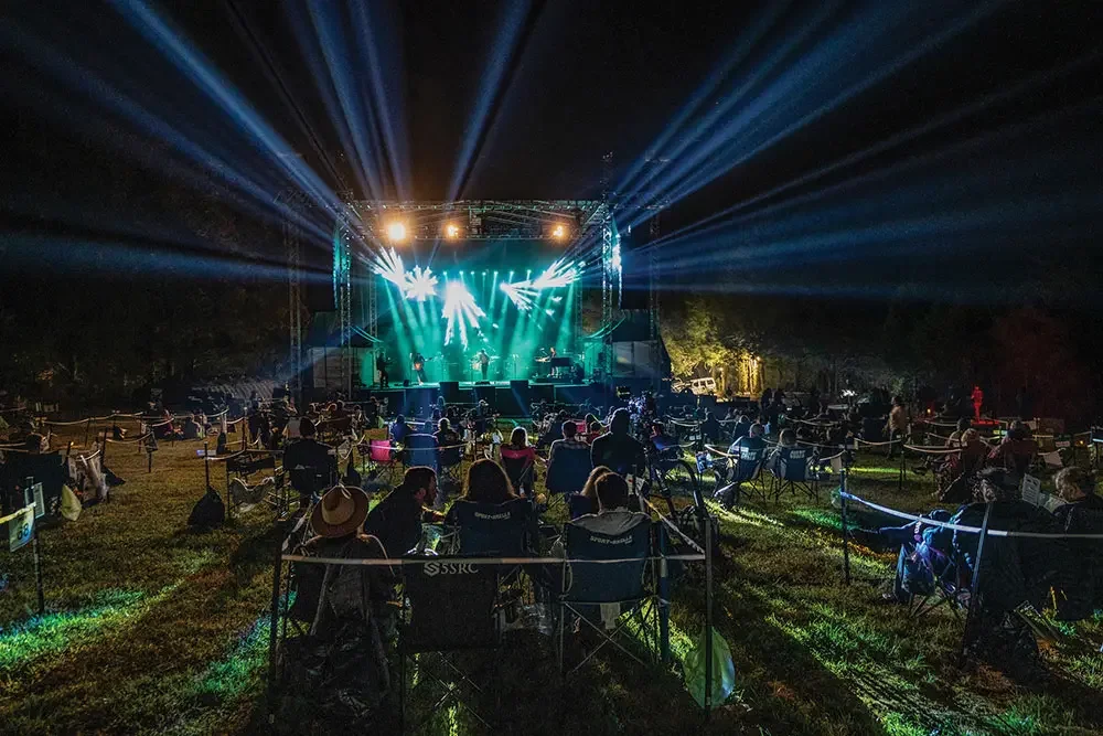 A lighted up outdoor stage is seen from the back of a concert crowd.