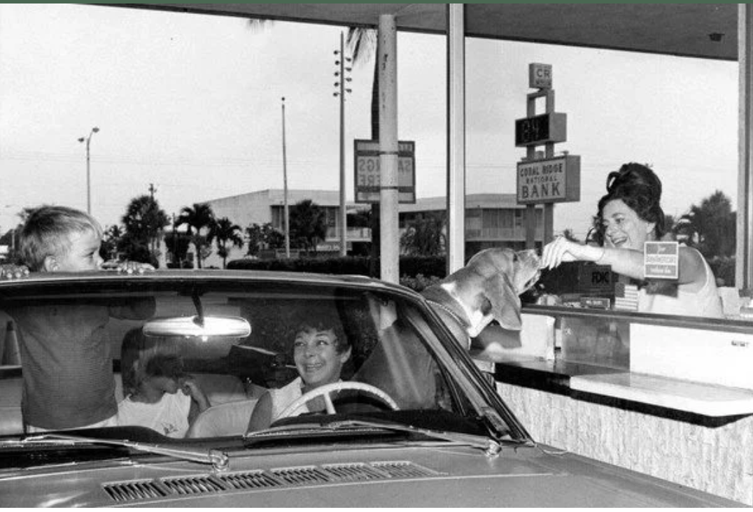 In a vintage black and white photo a server i a drive through window feeds a dog who is learning out of a car window.