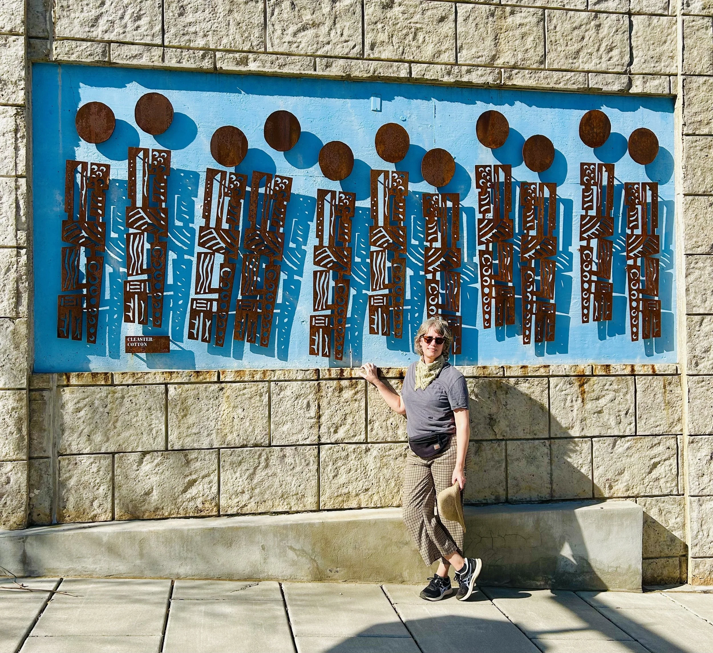 Alli Marshall wears a grey shirt, plaid pants, and gold bandana. She stands in front of a sculpture installation by the artist Cleaster Cotton.