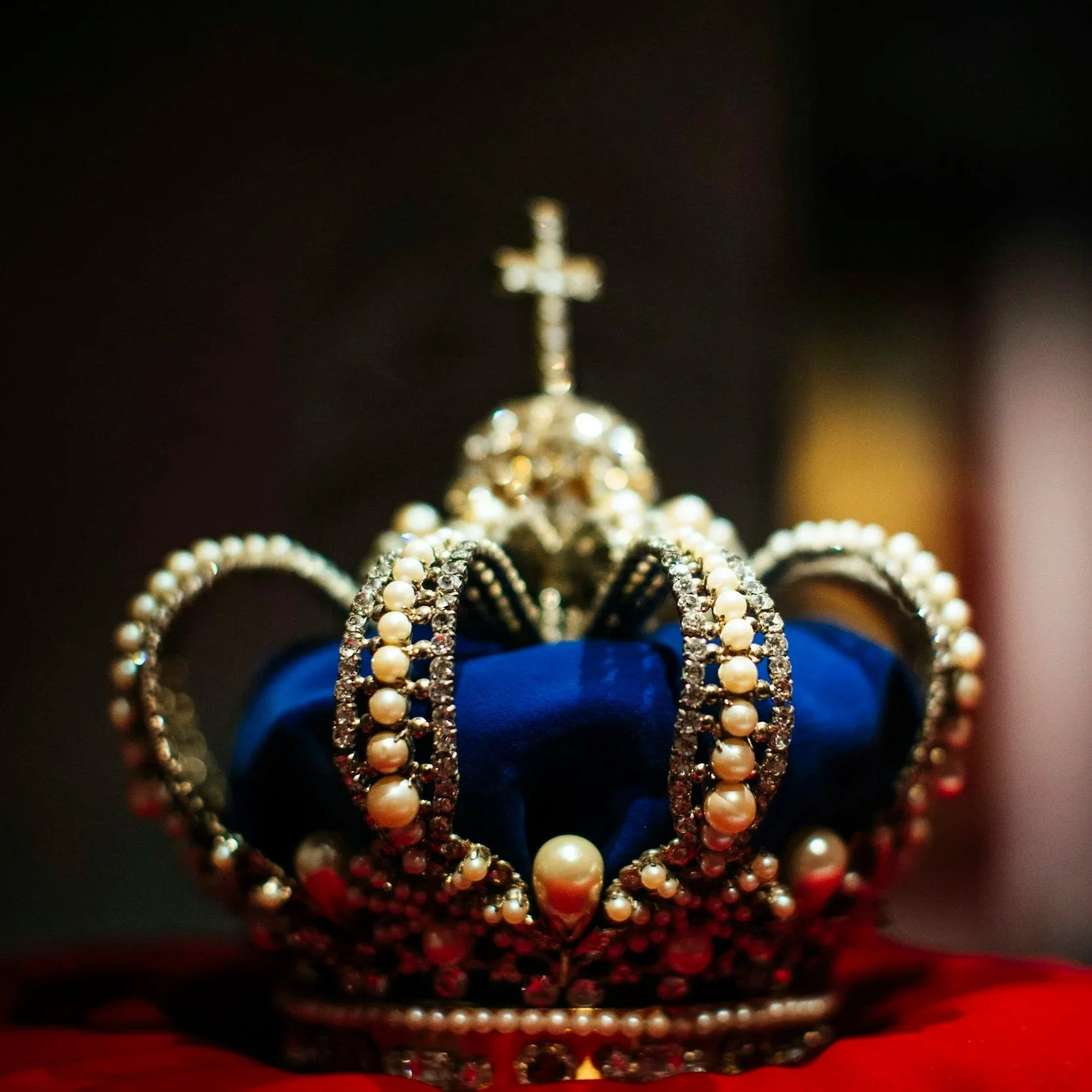 A jeweled royal crown adorned with pearls and rhinestones, with a small cross on top, resting on a red fabric surface.