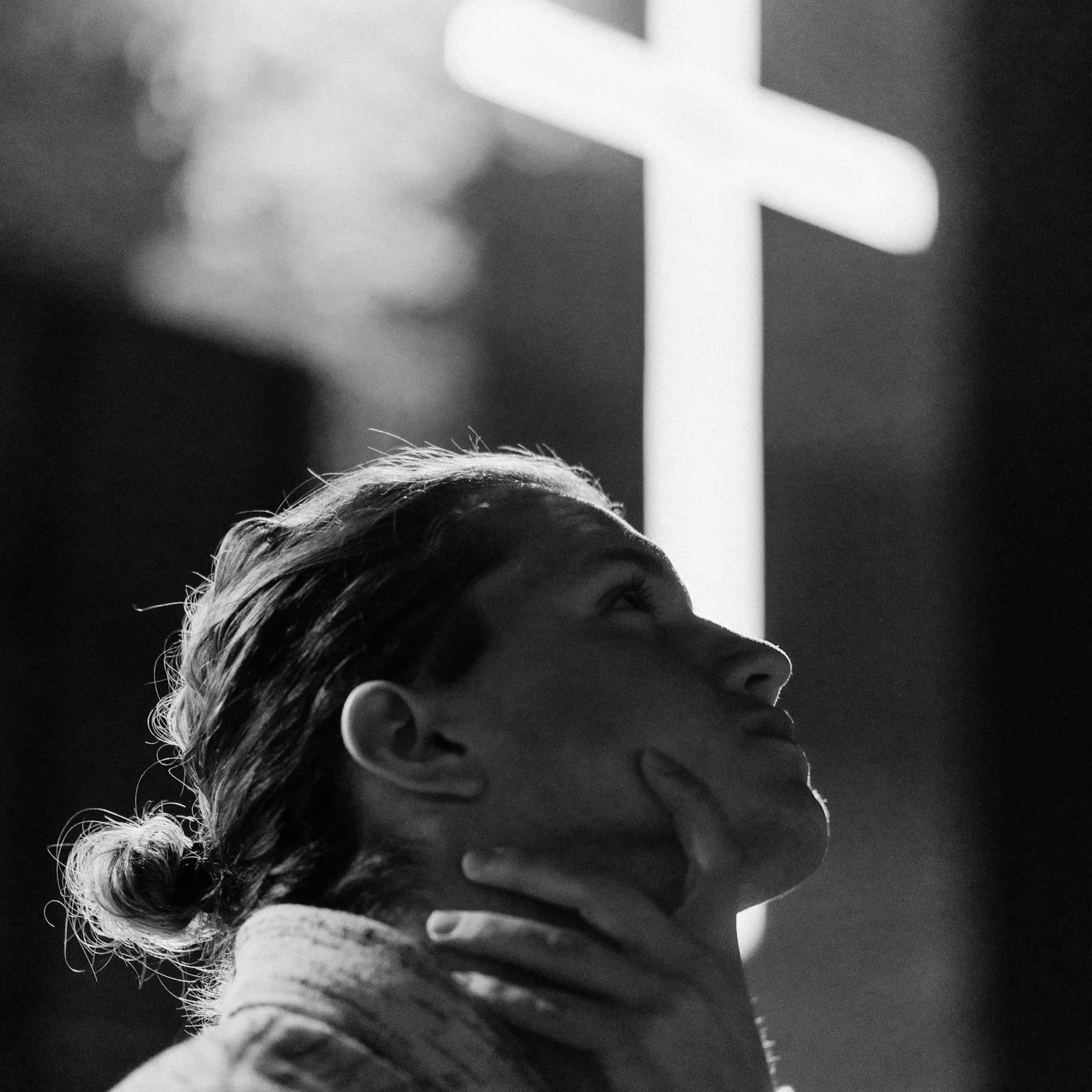 Black and white photo of a young woman with hair in a bun, looking up at a large illuminated cross on a dark background, with her hand on her neck.