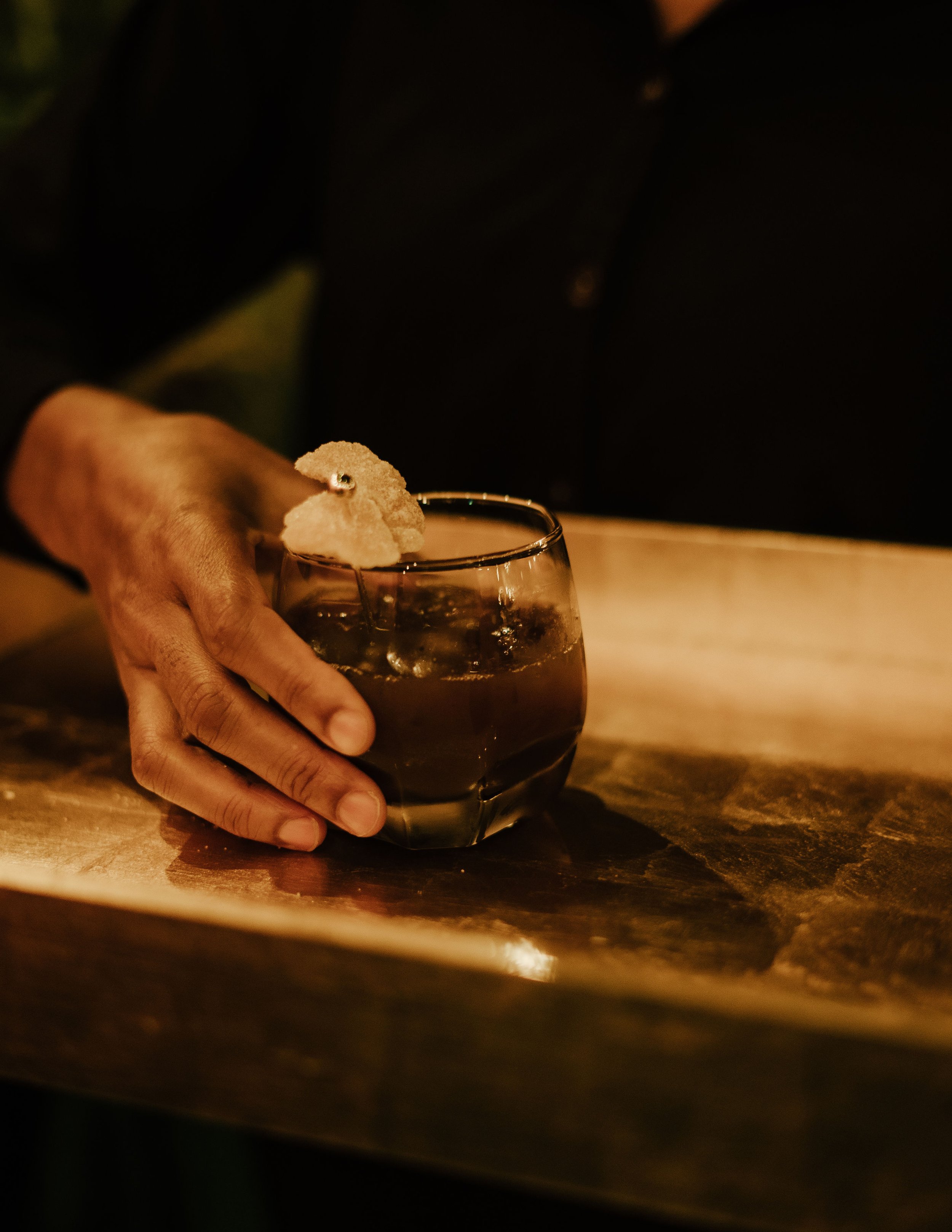 Hand holding a malt-based Apotheca mocktail garnished with some candied ginger in a gold lacquered rectangular tray.