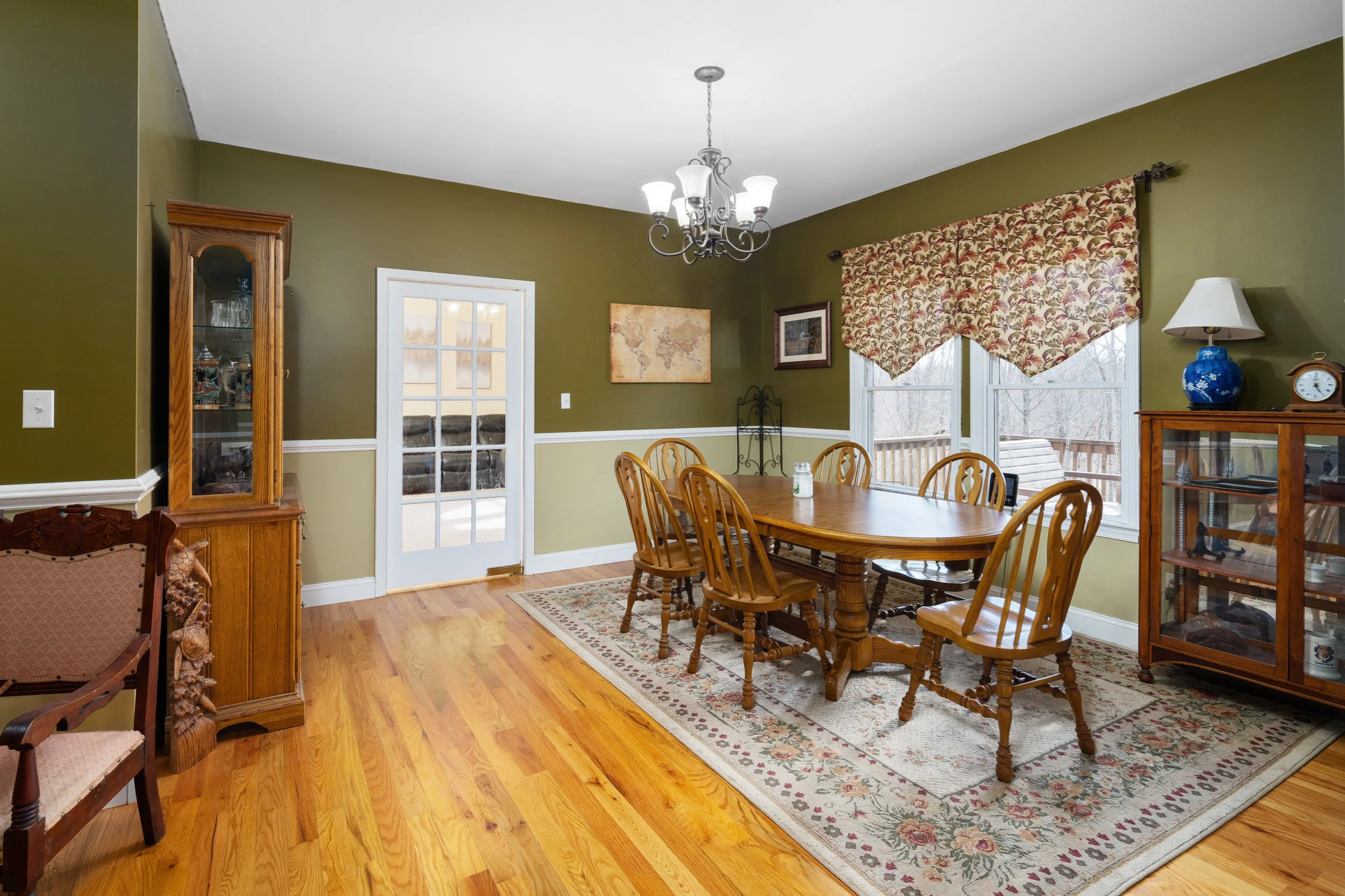 Dining room with green walls, wooden dining table, six wooden chairs, a patterned window valance, a glass-front cabinet with decorative items, a chandelier, an area rug, and a glass door leading to the outside deck.