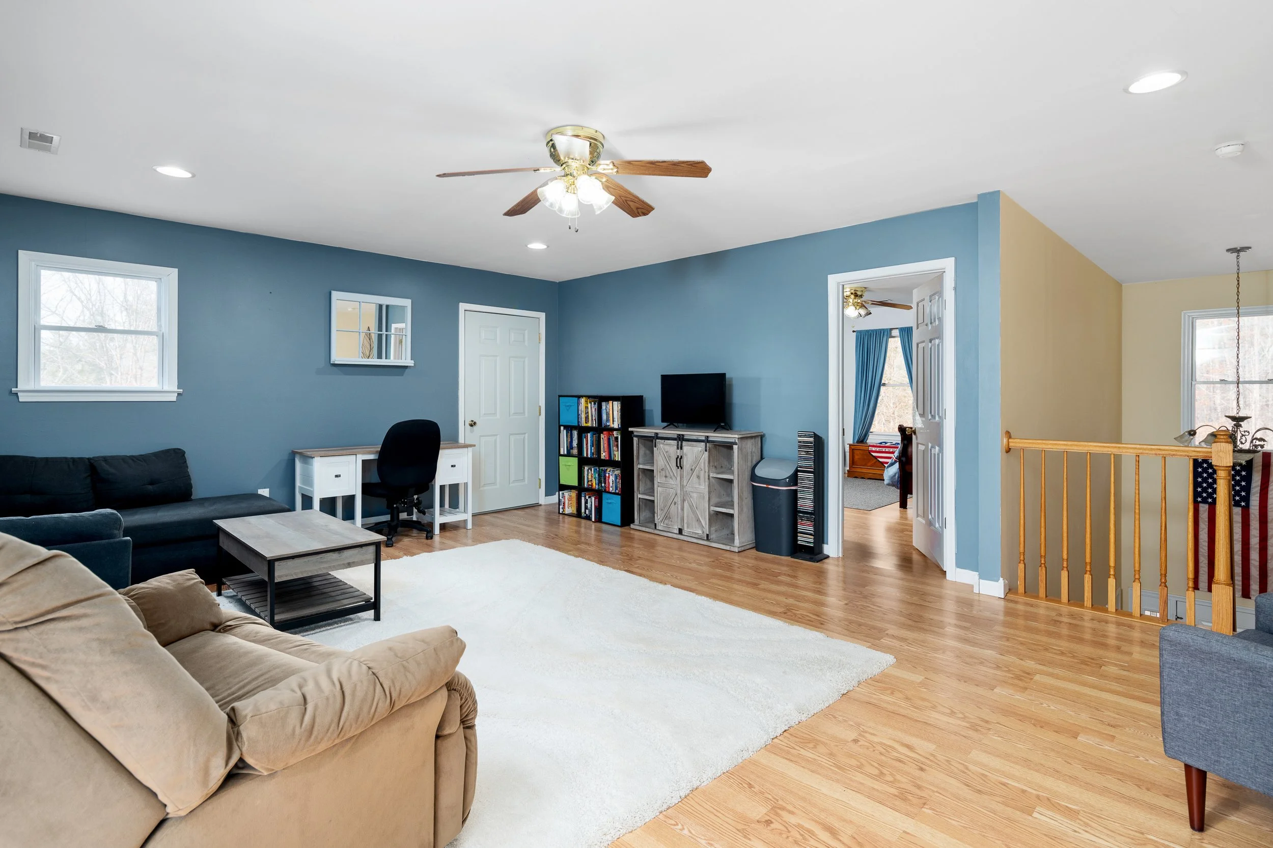 A spacious living room with blue and beige walls, hardwood floors, a ceiling fan, black and beige sofas, a white area rug, a small desk with a chair, a TV on a wooden stand, a bookshelf with books, a doorway with curtains, and a staircase with a wooden railing.