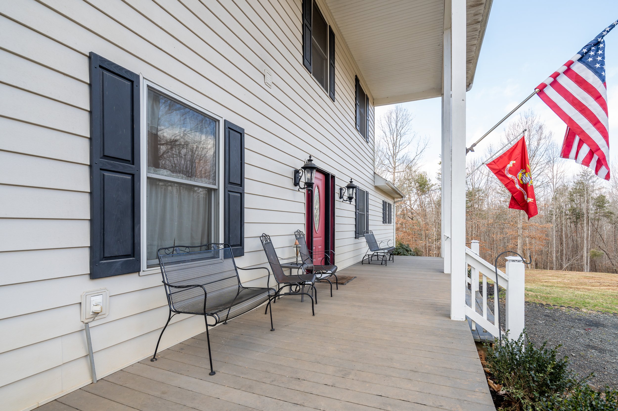 Front porch of a house with black shutters, a red door, and American and Marine flags, with outdoor seating and trees in the background.