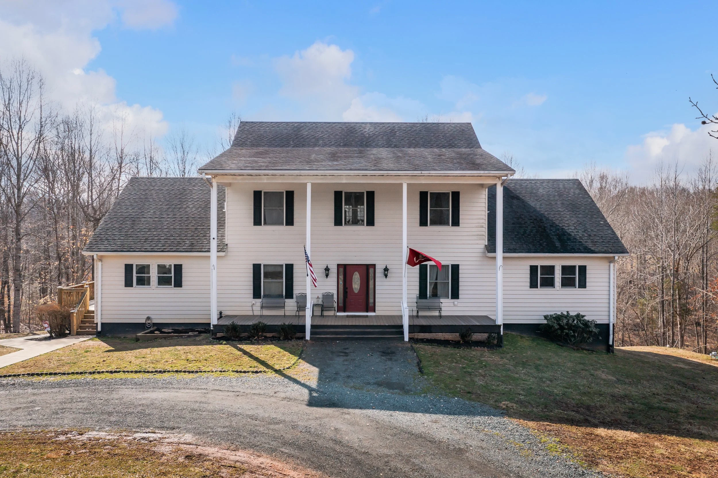 Front view of a two-story white house with black shutters and a red front door, with a porch supported by columns, a driveway leading up to it, and a wooded area in the background.