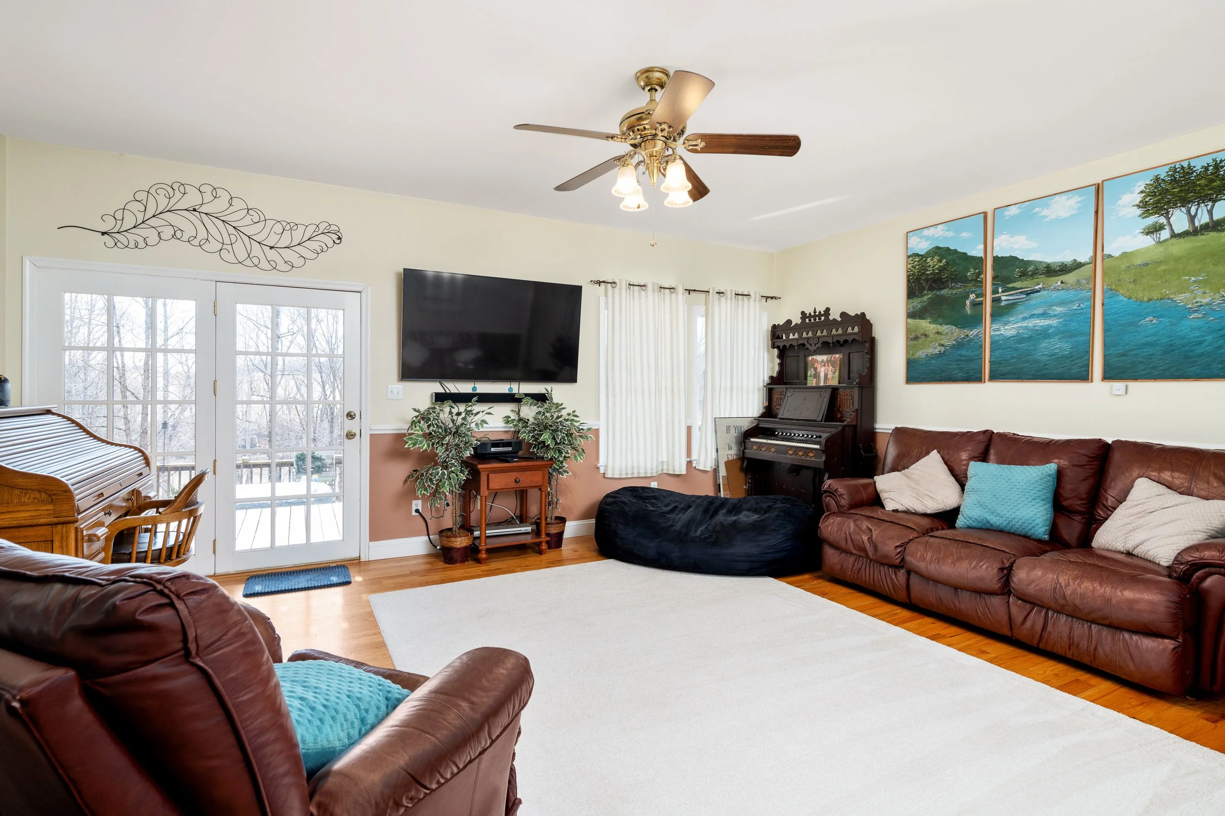 Living room with leather sofas, a large cream area rug, a flat-screen TV mounted on the wall, a wooden piano, a bean bag, and wall art depicting a river scene.