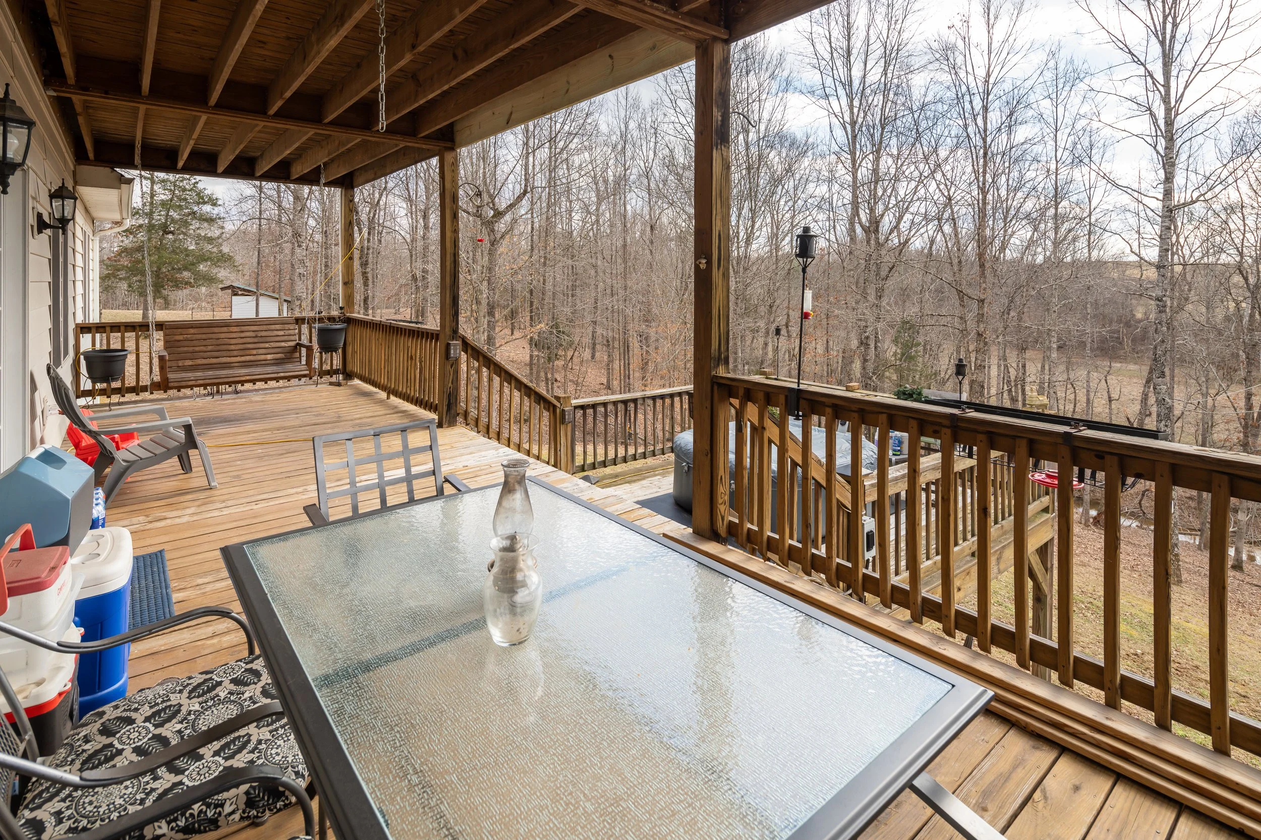 Wooden outdoor deck with glass table, chairs, swing, and hanging planters, overlooking a wooded backyard.
