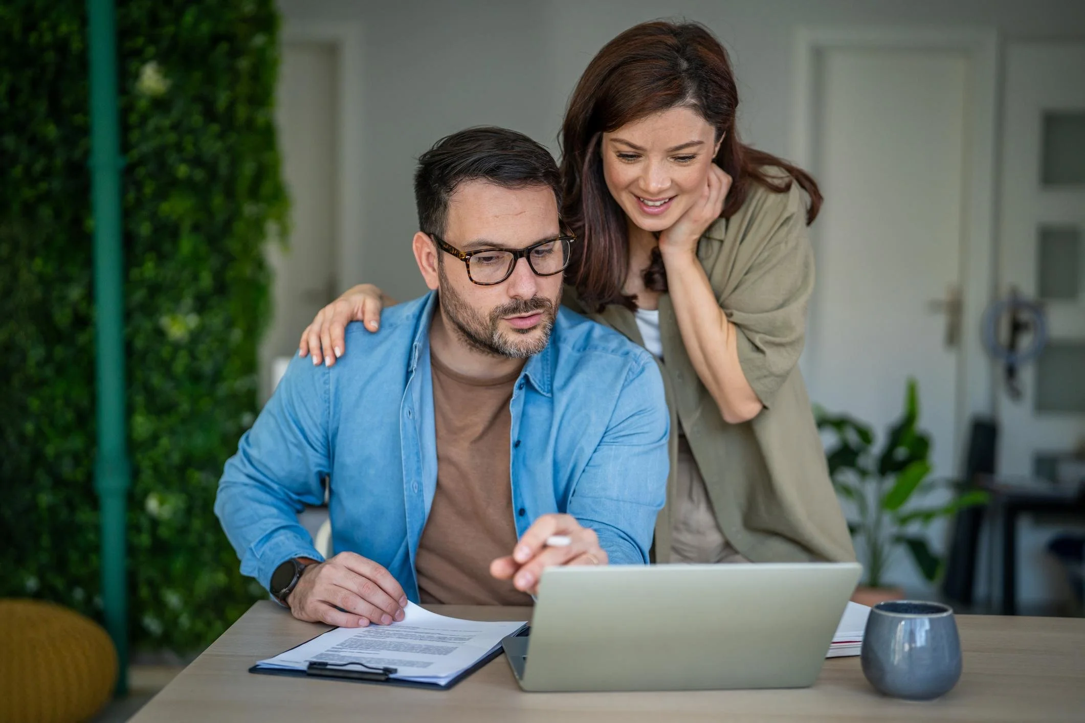 A man with glasses sitting at a desk looking at a laptop, with a woman standing beside him, resting her hand on his shoulder, both smiling and looking at the screen.
