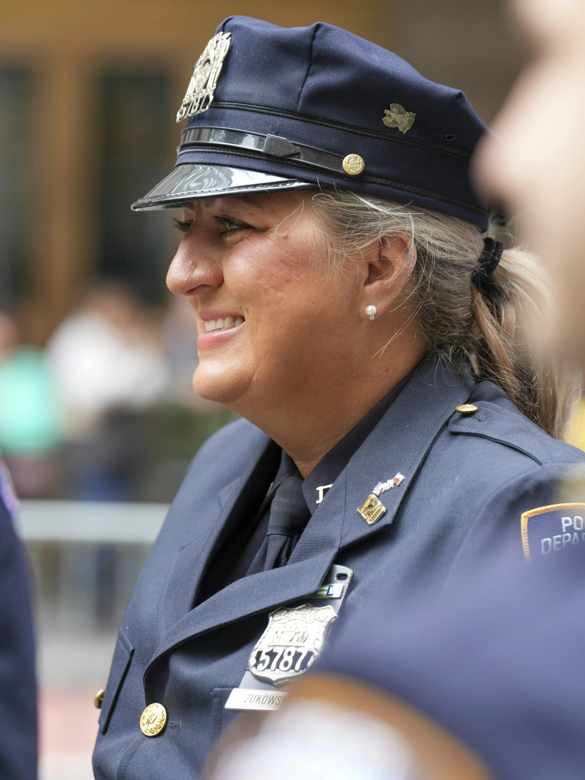 Close-up of a female police officer smiling, wearing a navy blue police uniform and hat, with badges and a pearl earring.