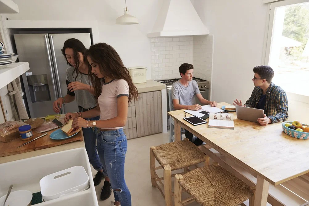 Four young people in a bright kitchen, two women preparing food at the counter and two men having a discussion at the table with a laptop and tablets.