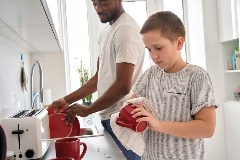 A man and a boy are washing dishes in a bright kitchen. The man is drying a plate, while the boy is holding a mug and a dish towel.
