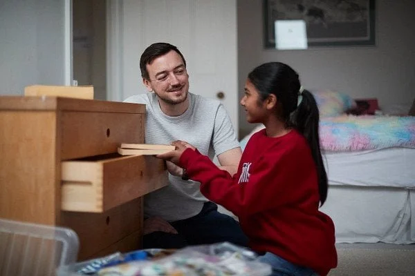 A young girl and an adult male are playing a card game at a wooden dresser in a bedroom.