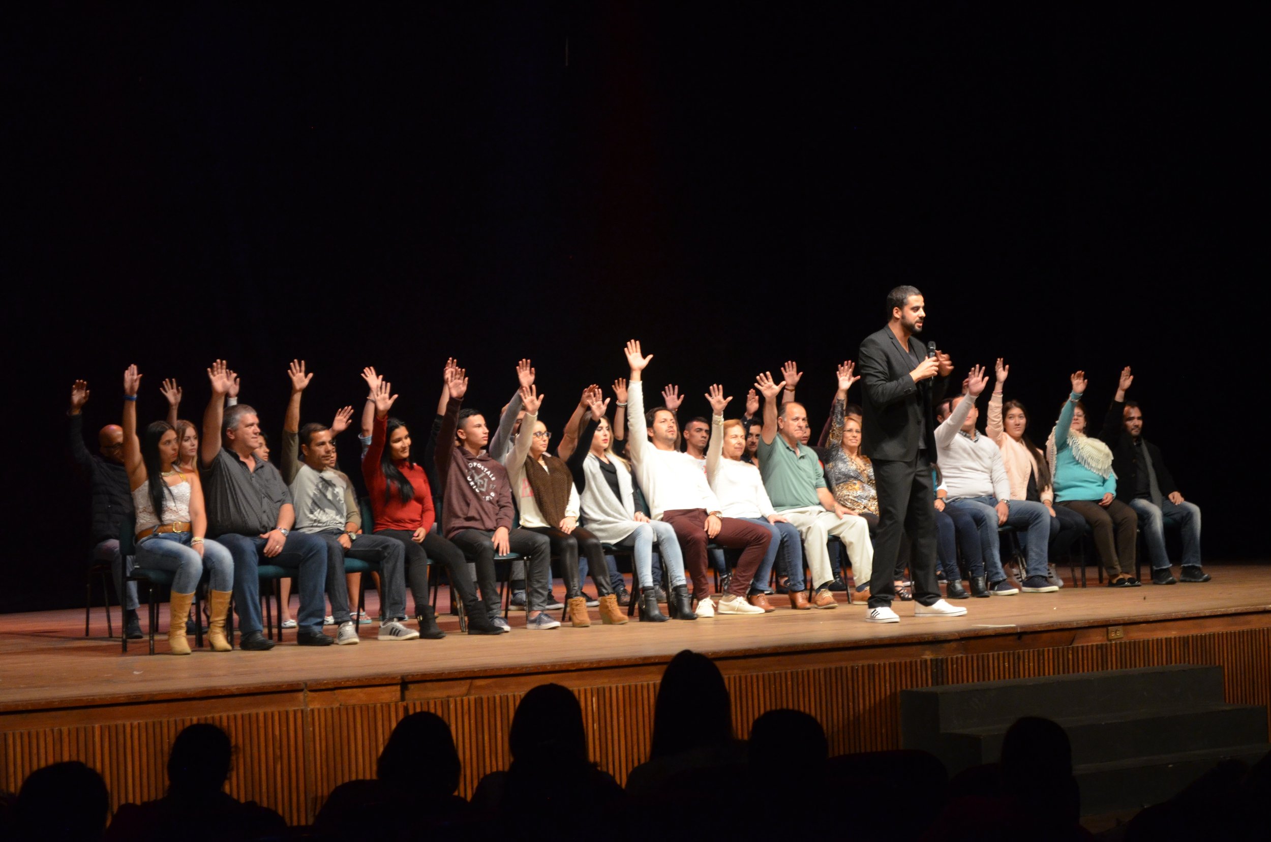Grupo de personas en un escenario, algunas se ven descansando o dormidas, mientras un hombre con micrófono de pie parece estar hablando.