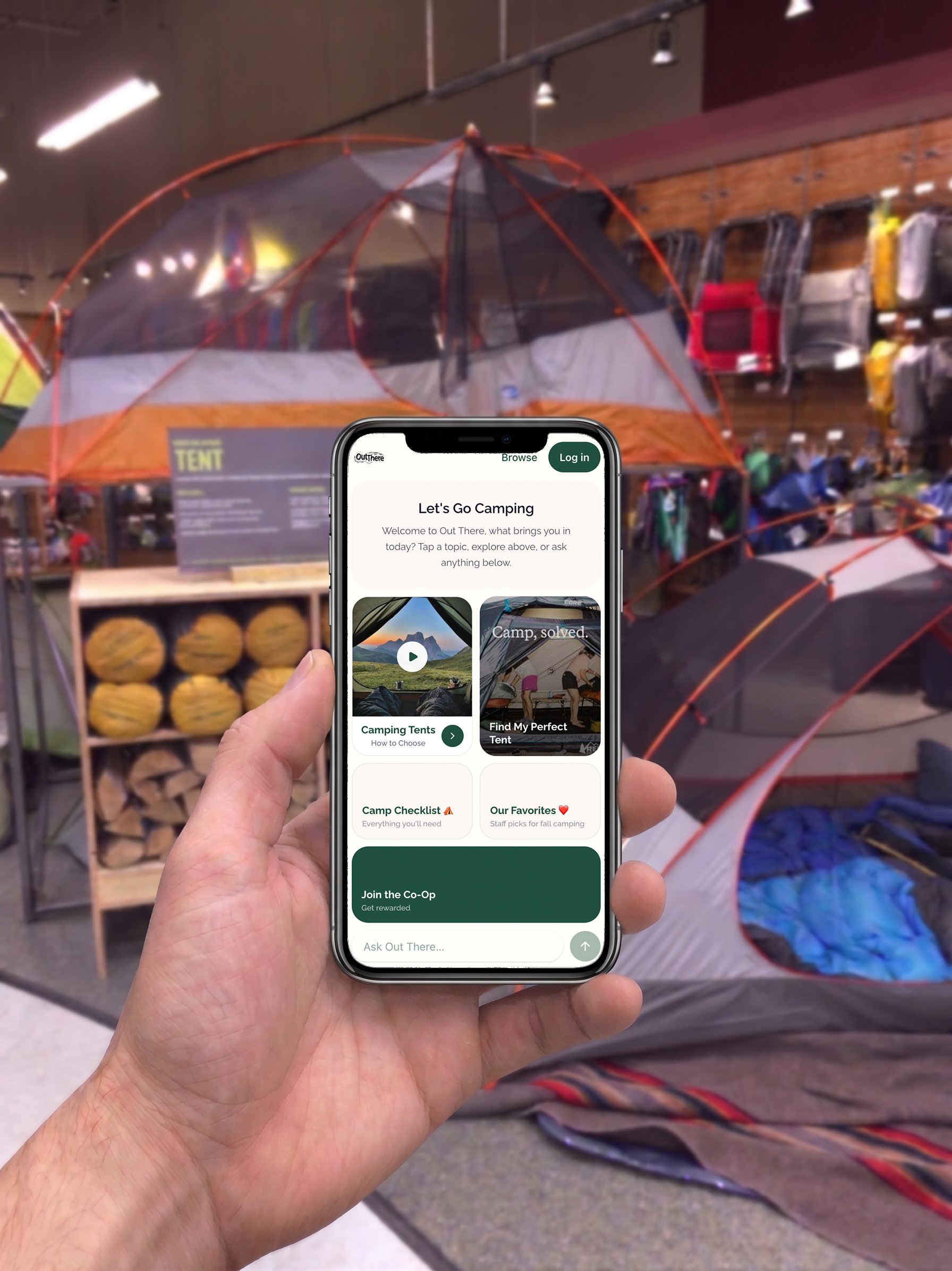 A person holding a smartphone displaying a camping app in front of a retail display of camping supplies, including a large outdoor tent, logs, and camping gear.