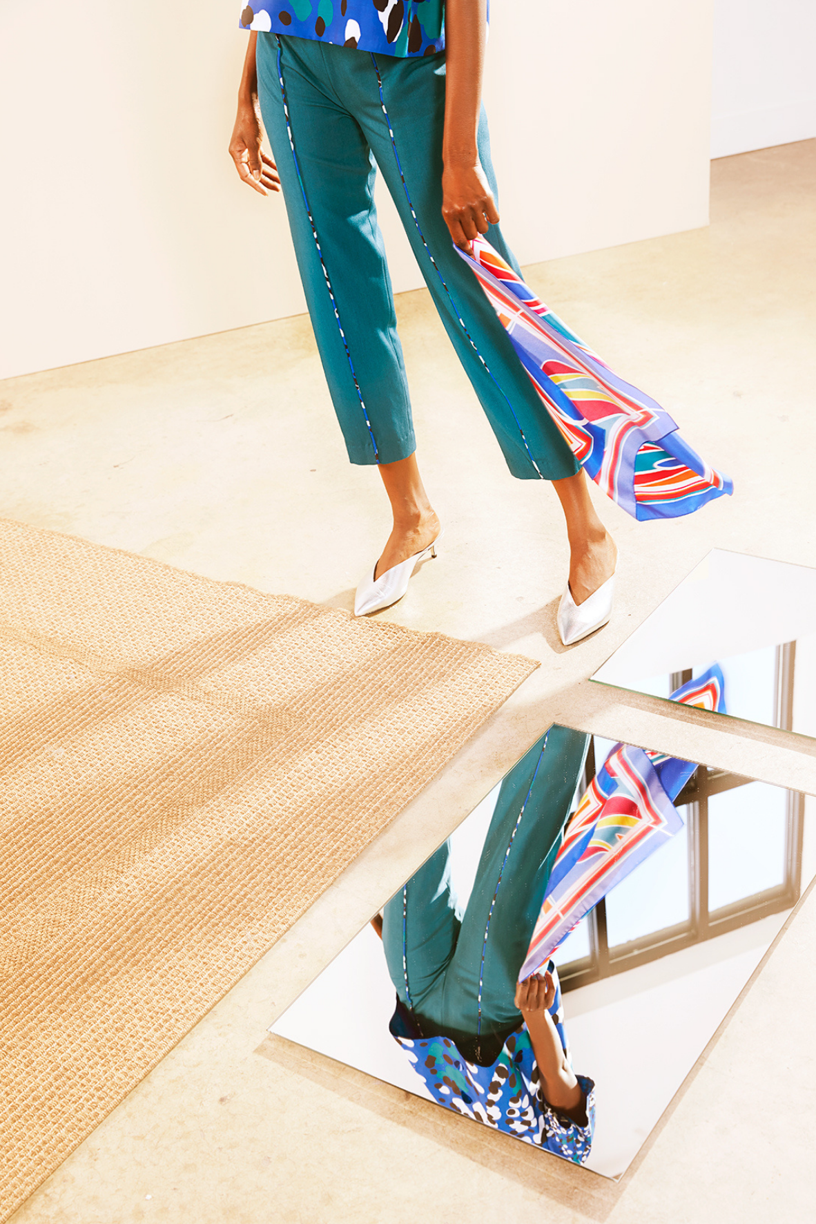 Woman wearing teal pants and a patterned top standing near mirrors on a beige floor with a beige rug nearby.